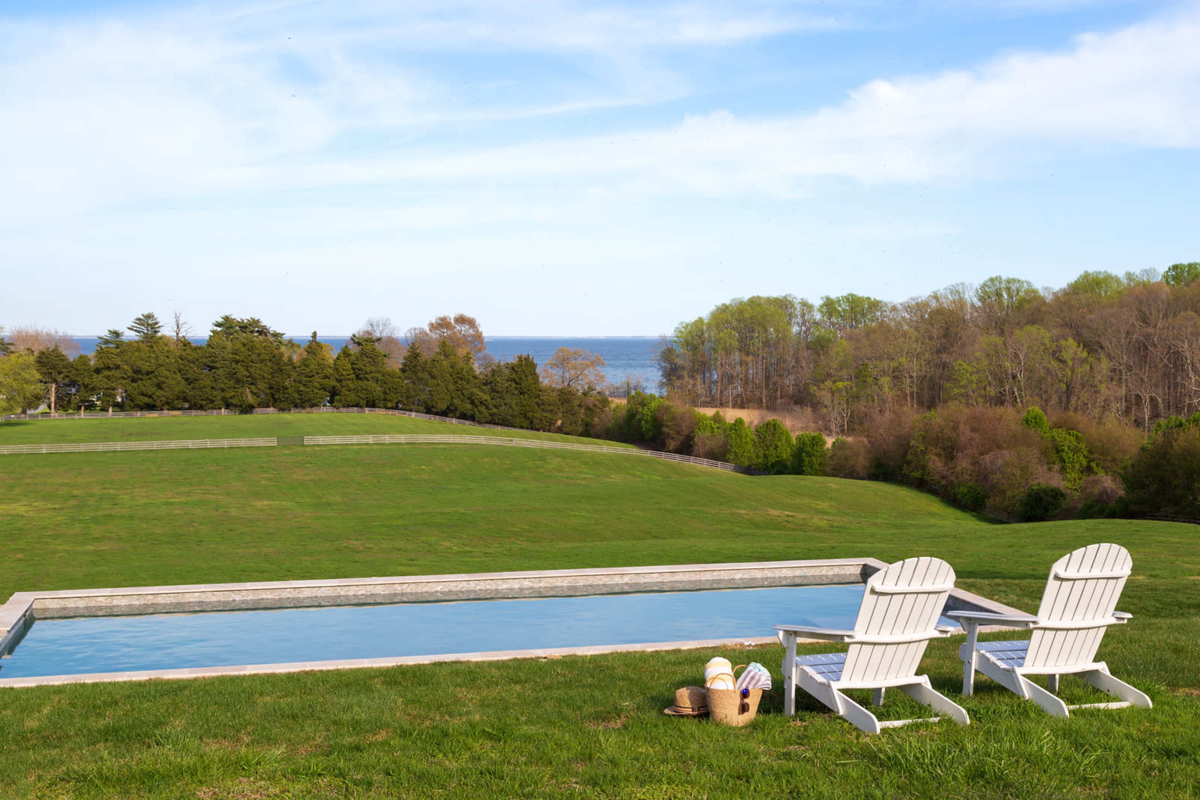 Two white Adirondack chairs sit beside a long, narrow swimming pool overlooking a grassy field and a body of water in the distance.