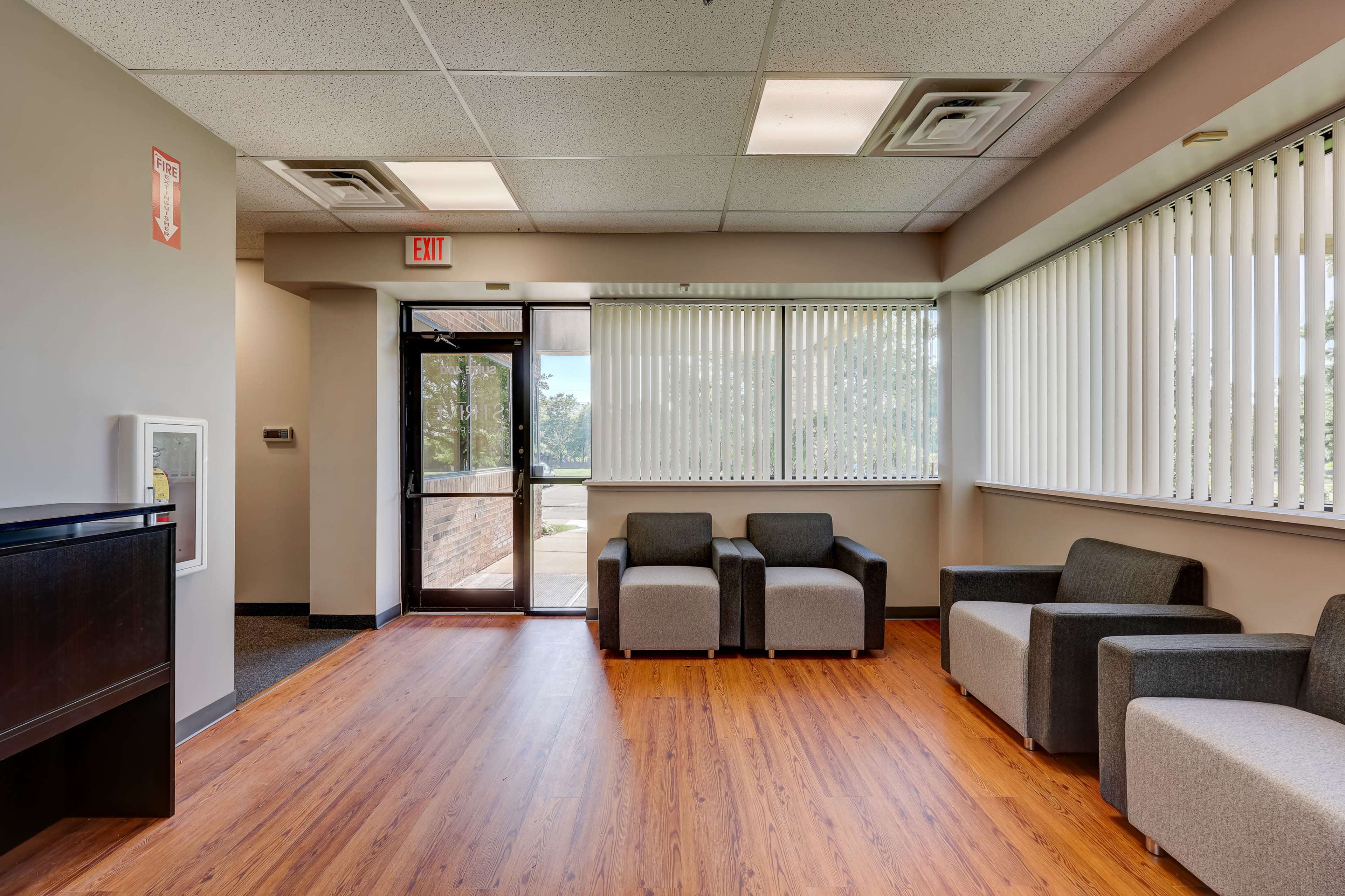 A waiting area with gray armchairs, wooden flooring, large windows with blinds, and an entrance door.