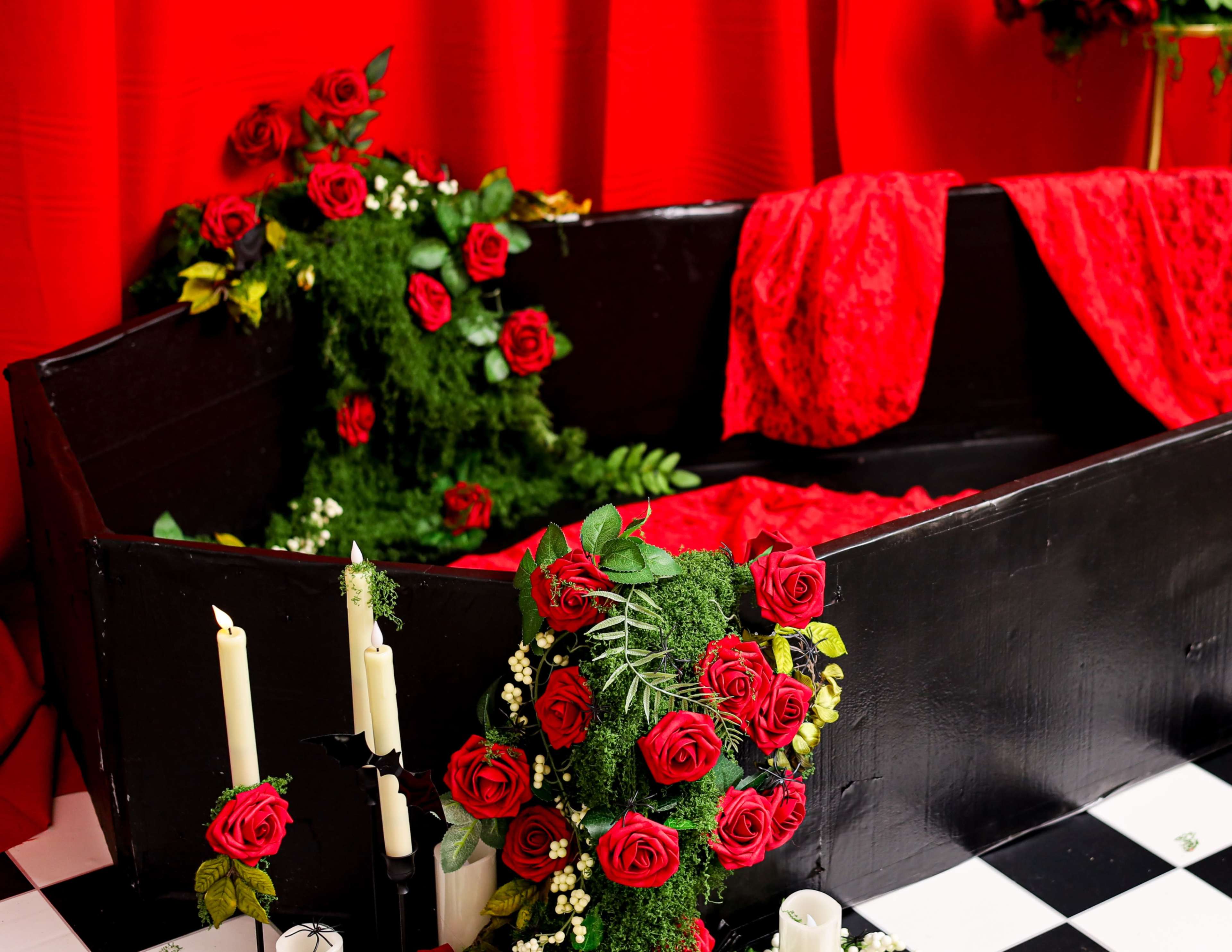 A black coffin is adorned with red roses and greenery, surrounded by candles, against a vibrant red backdrop.