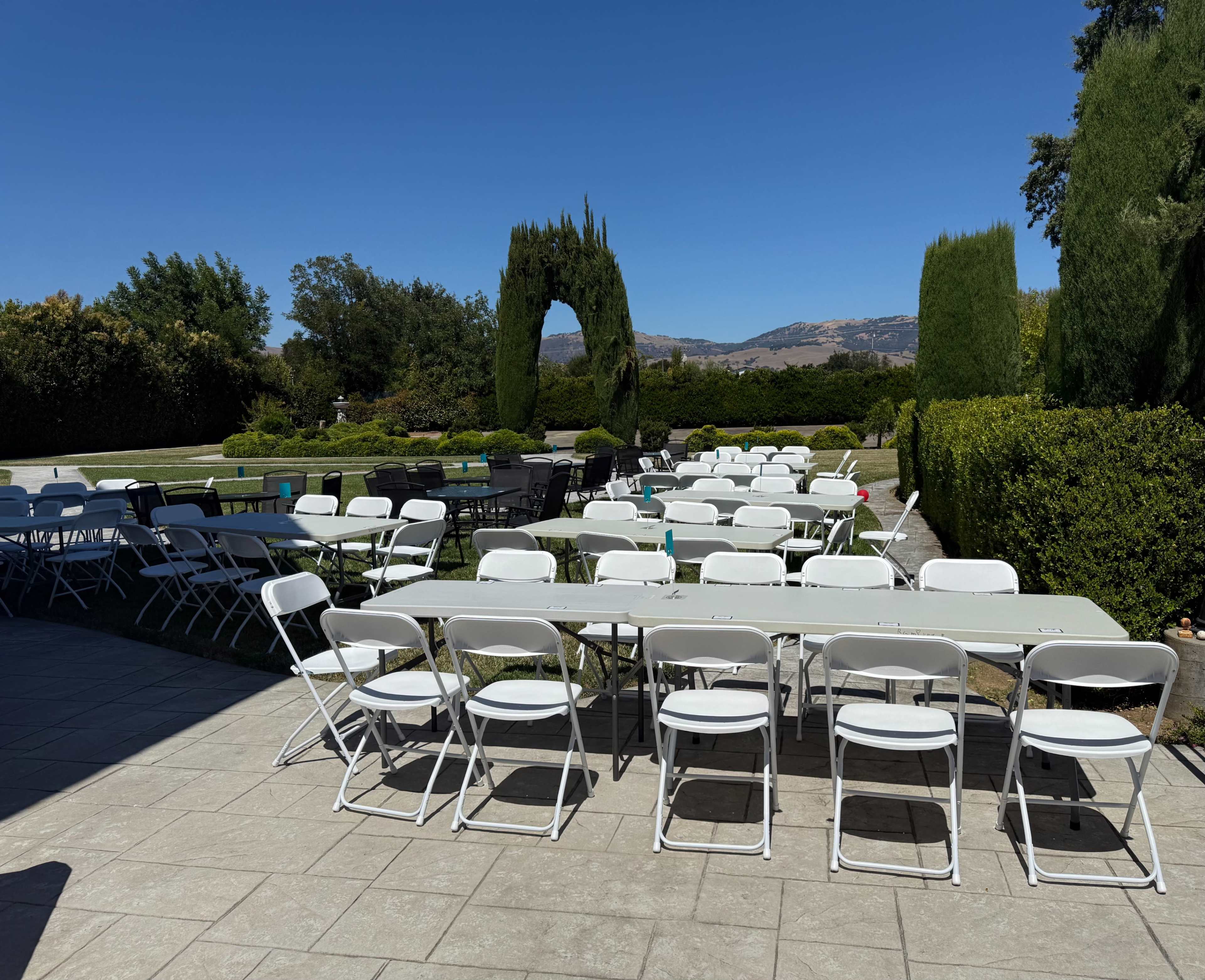 A setup of white folding tables and chairs is arranged outdoors in a garden with neatly trimmed hedges under a clear blue sky.