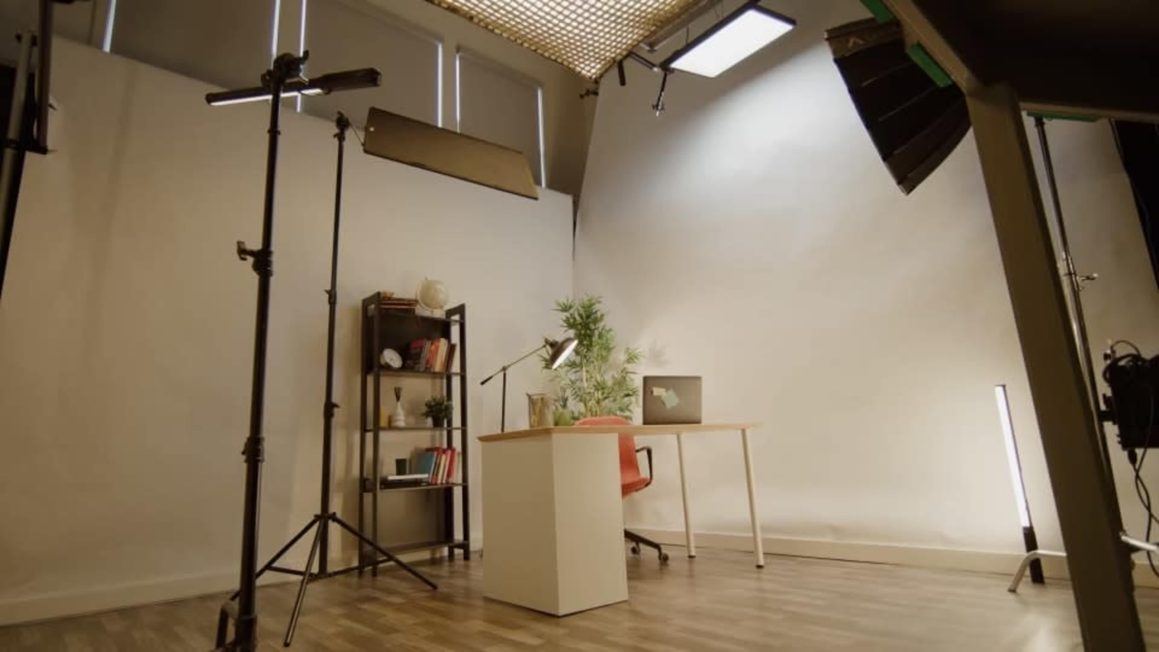 The image shows a minimalist studio setup with a desk, chair, shelving unit, and lighting equipment against a plain backdrop.