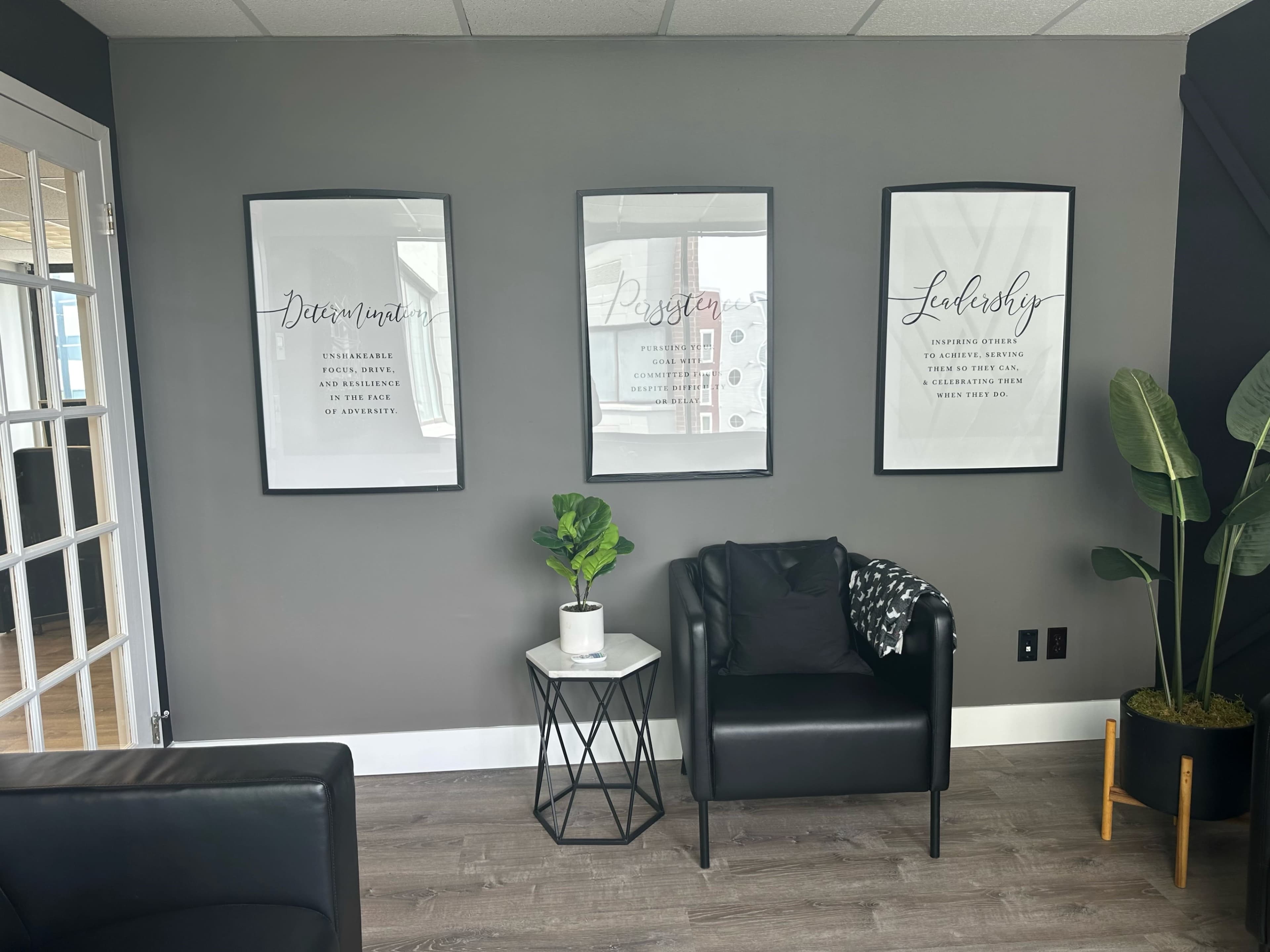 A modern office space featuring three framed motivational prints on a gray wall, a potted plant on a side table, and a black armchair.