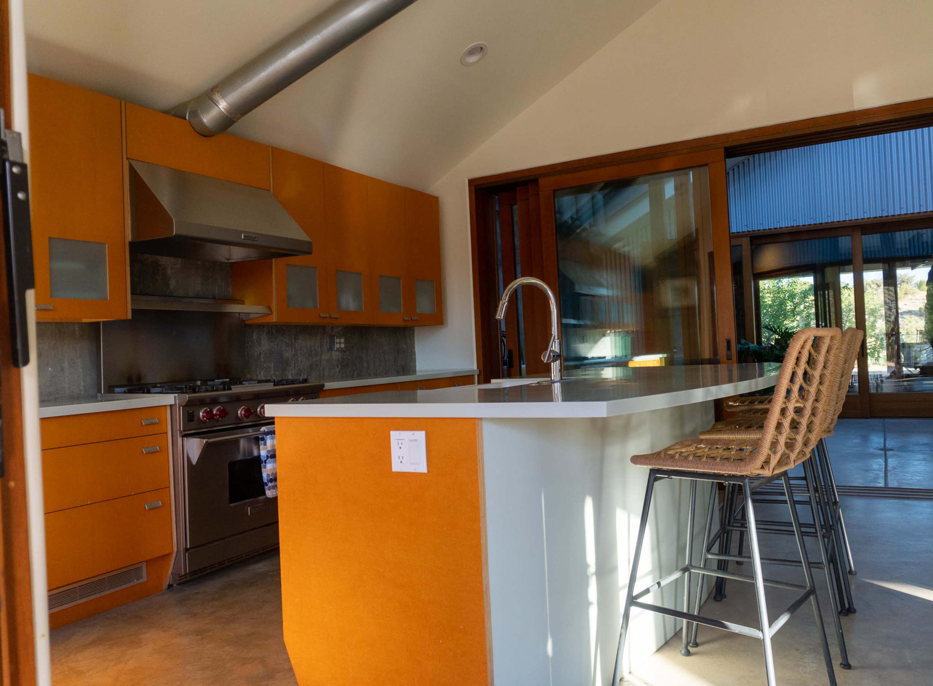 A modern kitchen with orange cabinetry, stainless steel appliances, and bar stools at a white countertop.