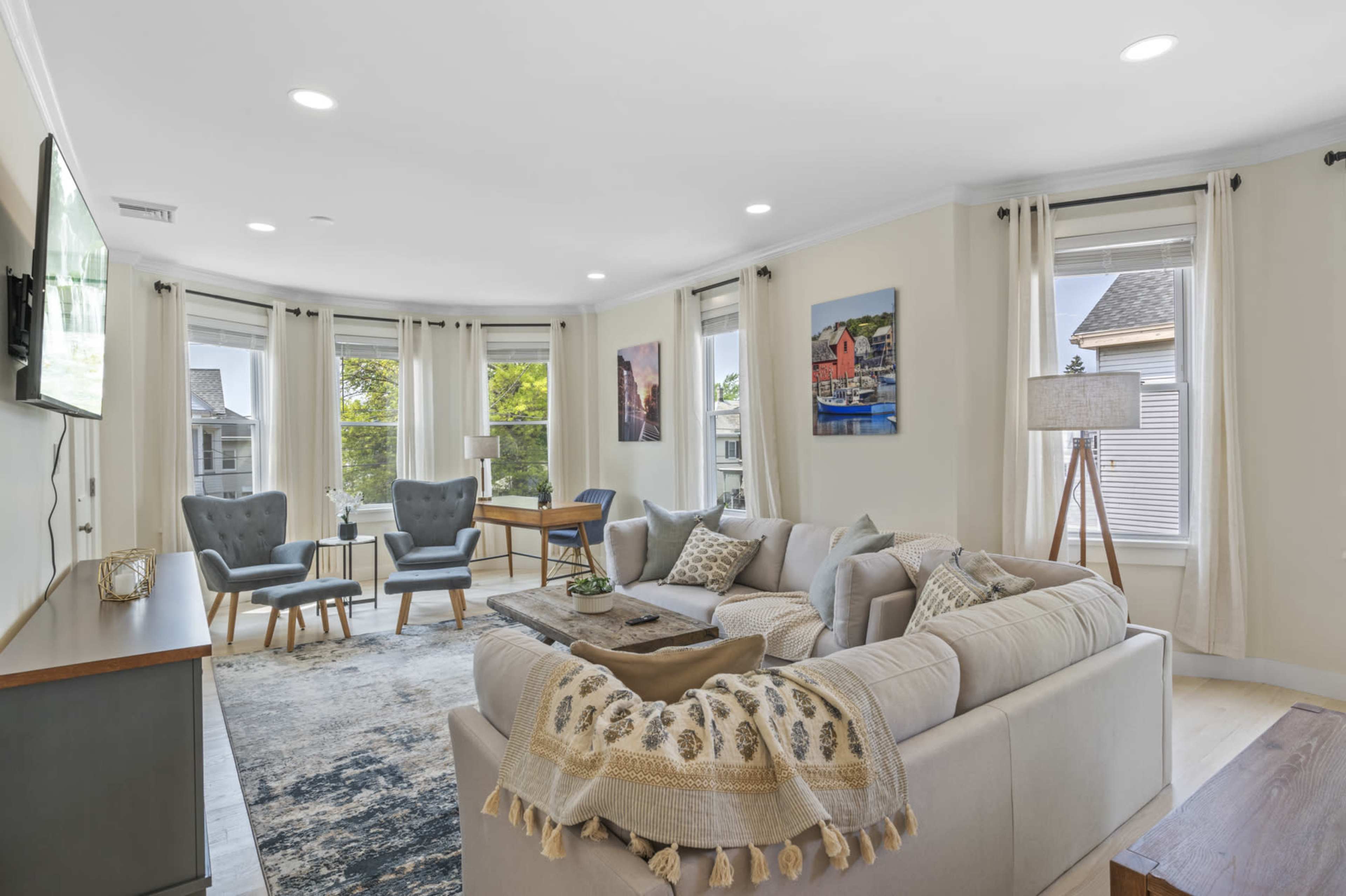 A modern living room features a large sectional sofa, two armchairs, and a coffee table, with large windows providing natural light.