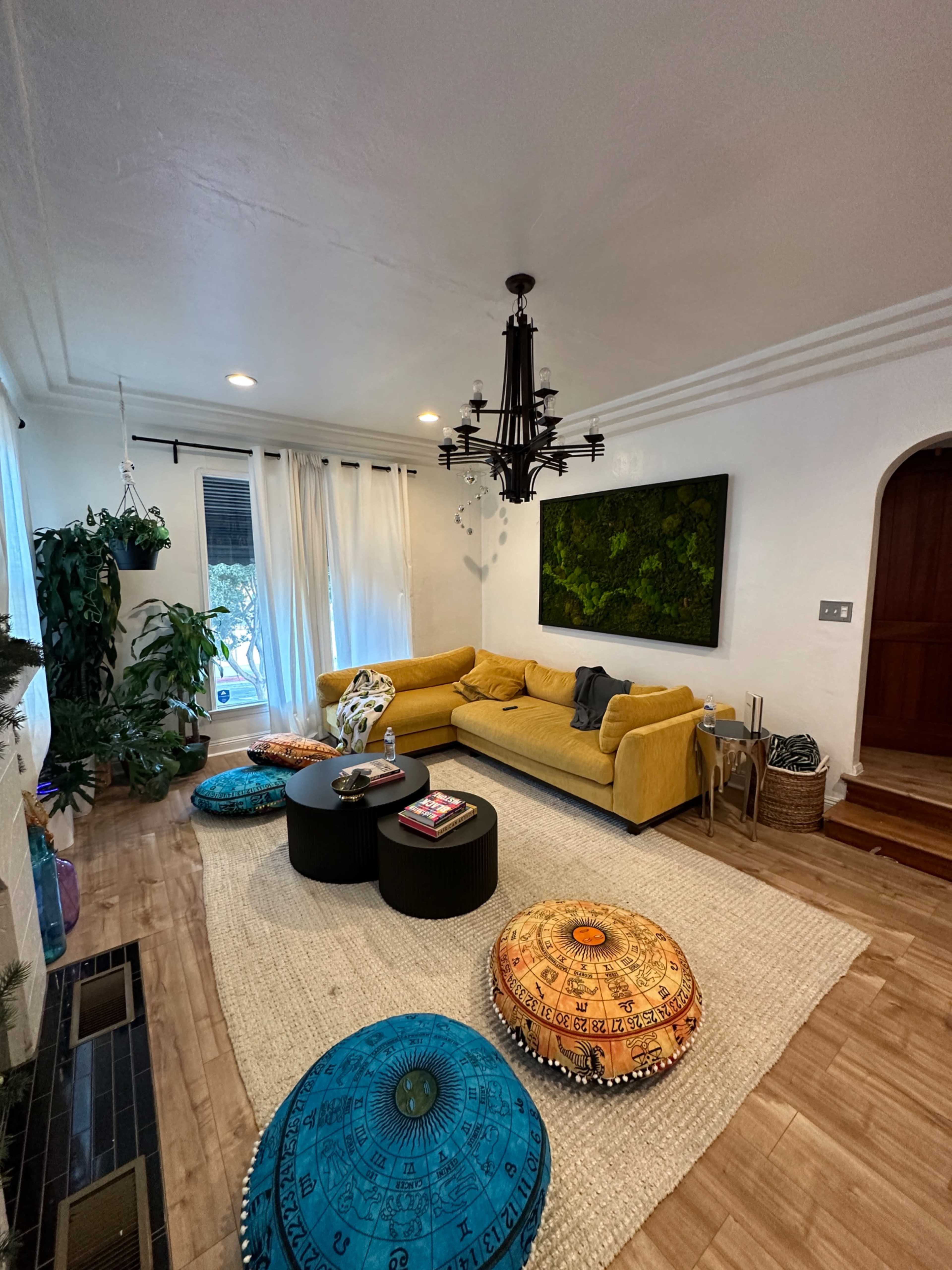 The living room features a yellow sofa, two black circular tables, decorative poufs, and a large wall-mounted art piece, with plants and natural light coming through the windows.