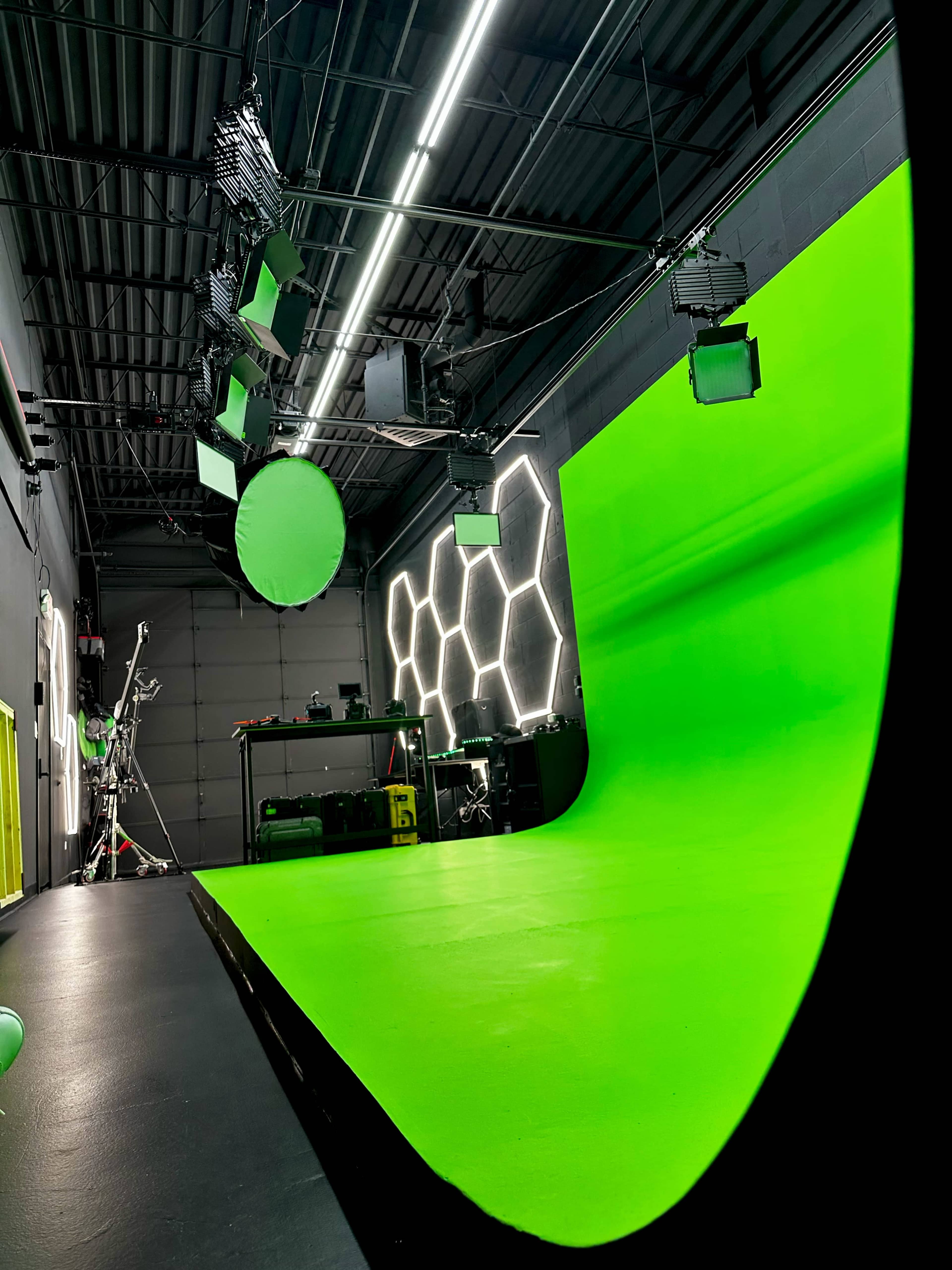 The image shows a green screen studio with a large green backdrop, various lights hanging from the ceiling, and equipment arranged on a table in the background.