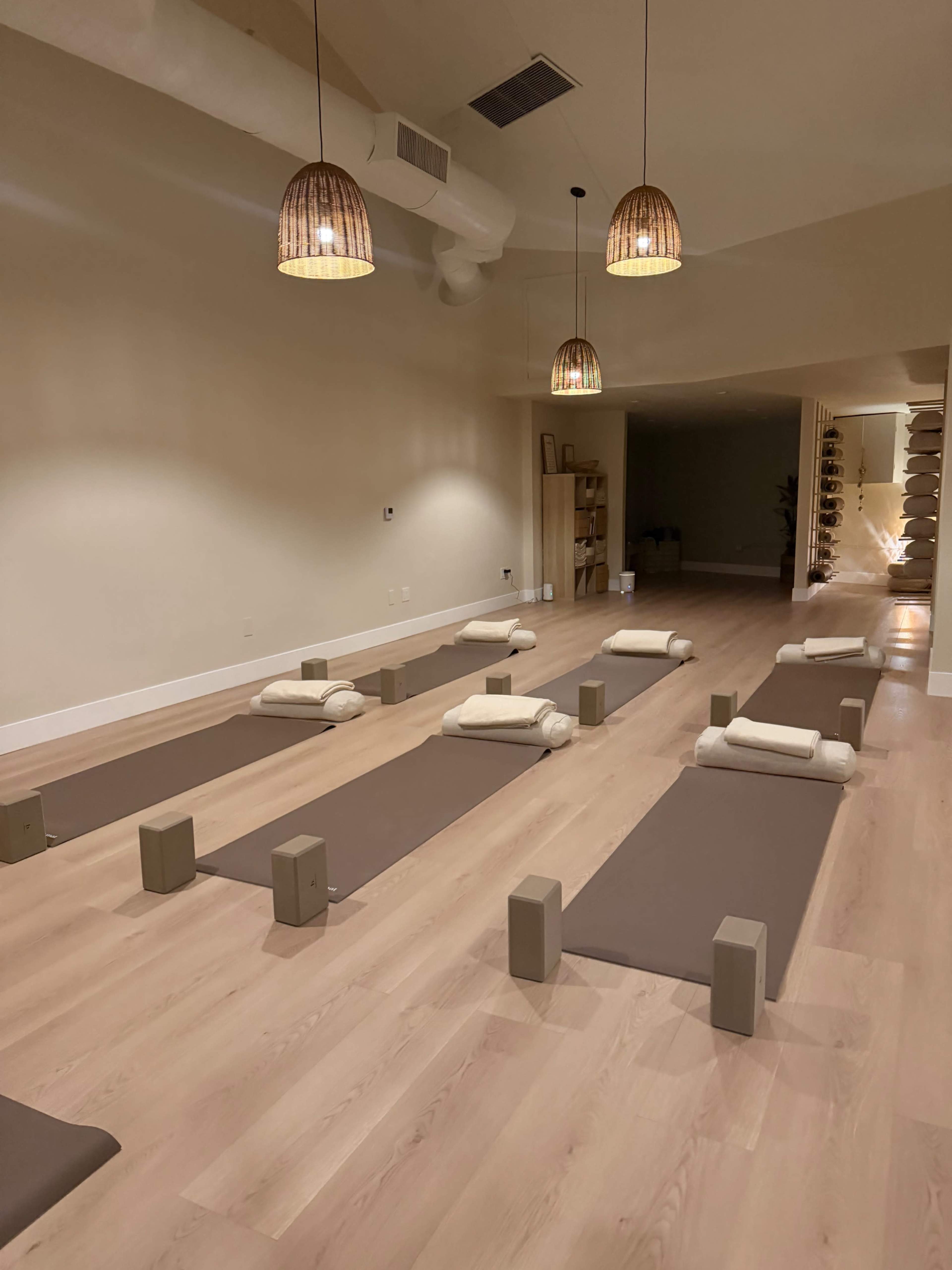 The image shows a serene yoga studio with multiple yoga mats arranged neatly on a light wooden floor, each paired with props for practice.