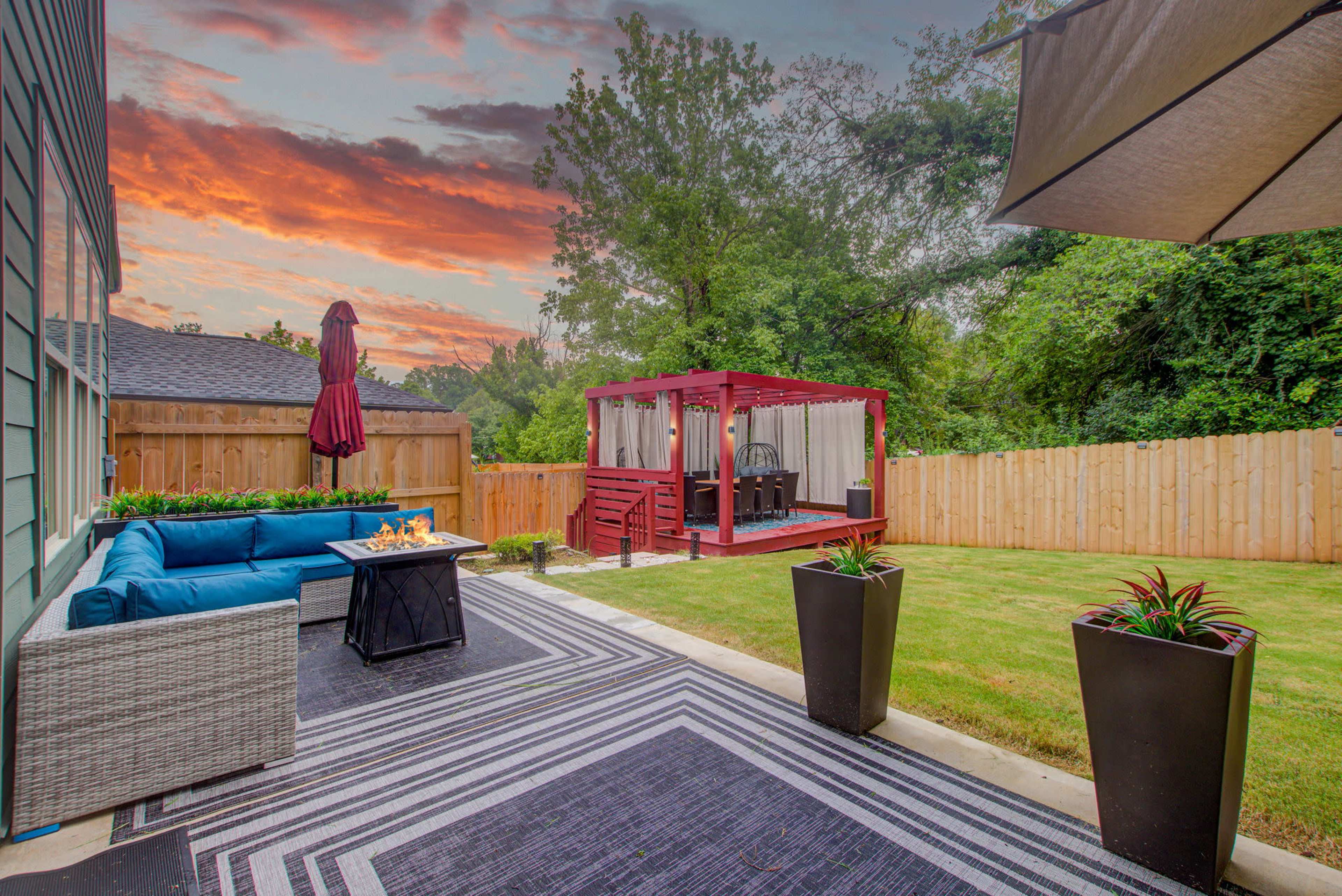 A landscaped backyard featuring a blue outdoor sofa, a wooden gazebo, and a vibrant sunset sky.