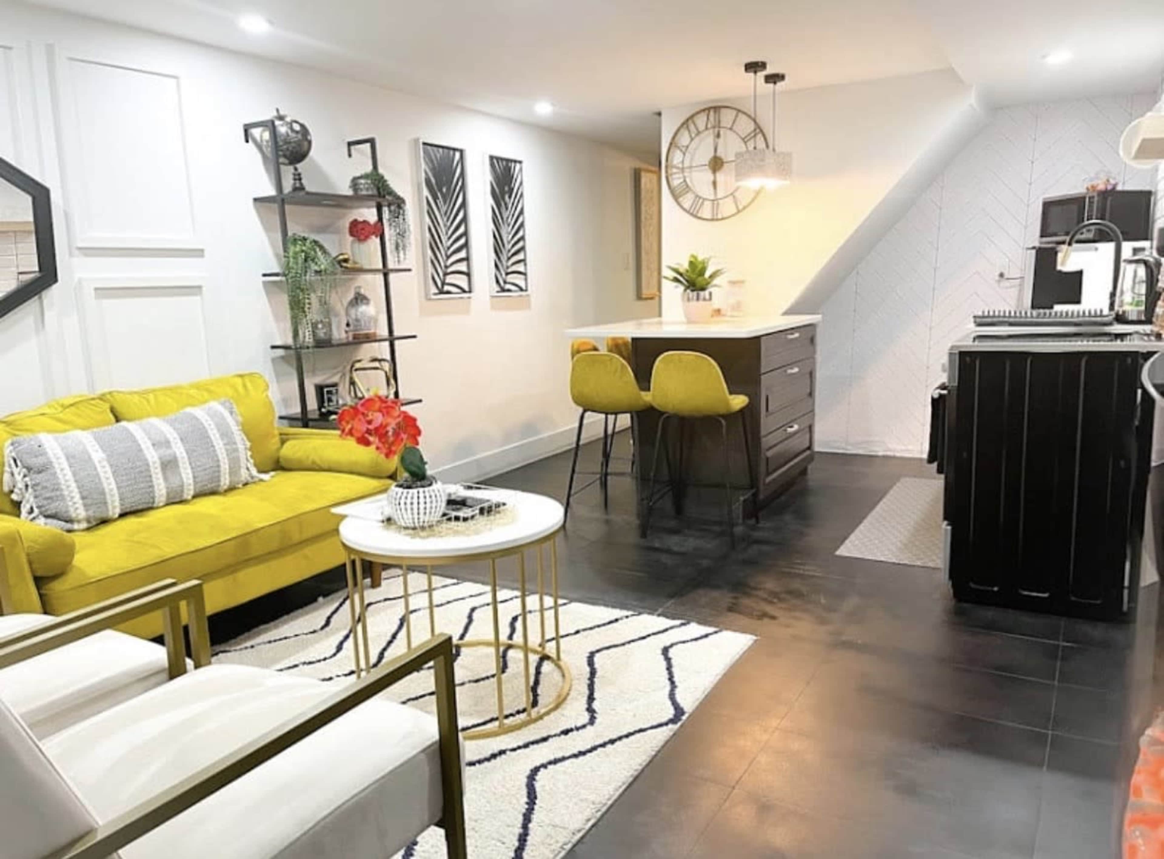 The image shows a modern living area with a yellow sofa, a round coffee table, and a dining area featuring two yellow chairs and a kitchen in the background.