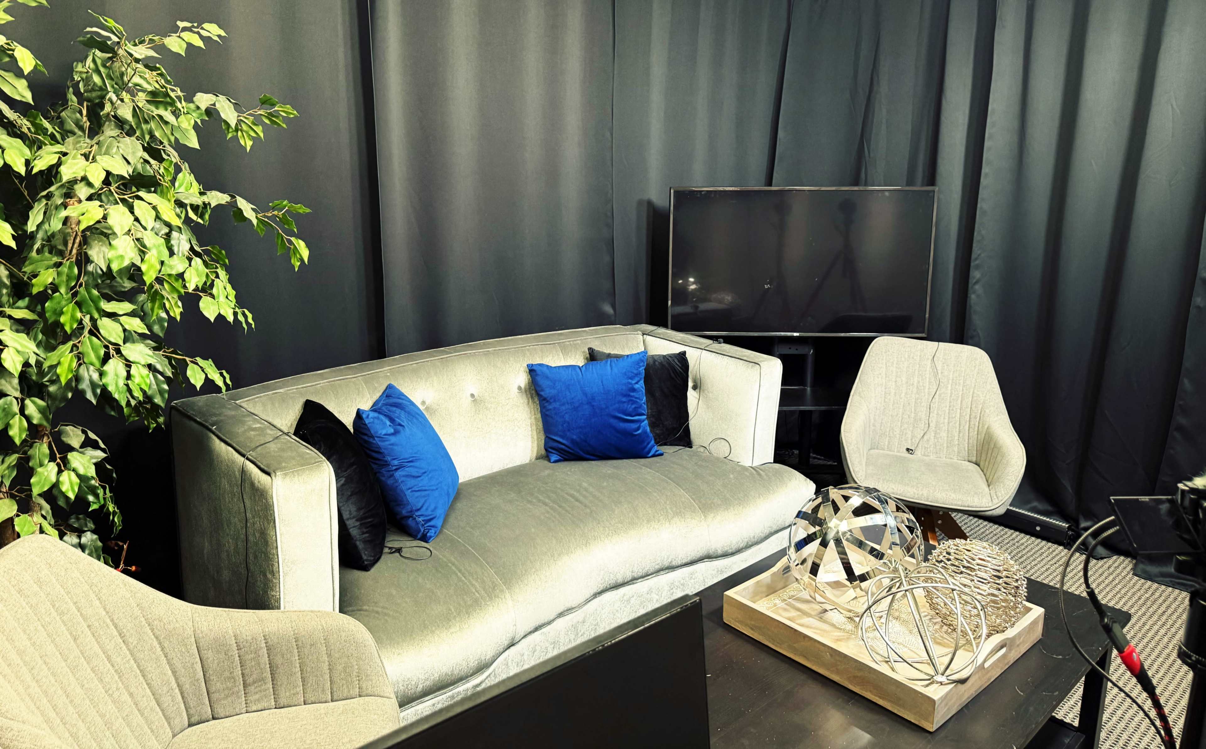 A cozy seating area features a light gray couch adorned with blue pillows, accompanied by two light-colored chairs and a decorative table with a sculpture, against a backdrop of black curtains.