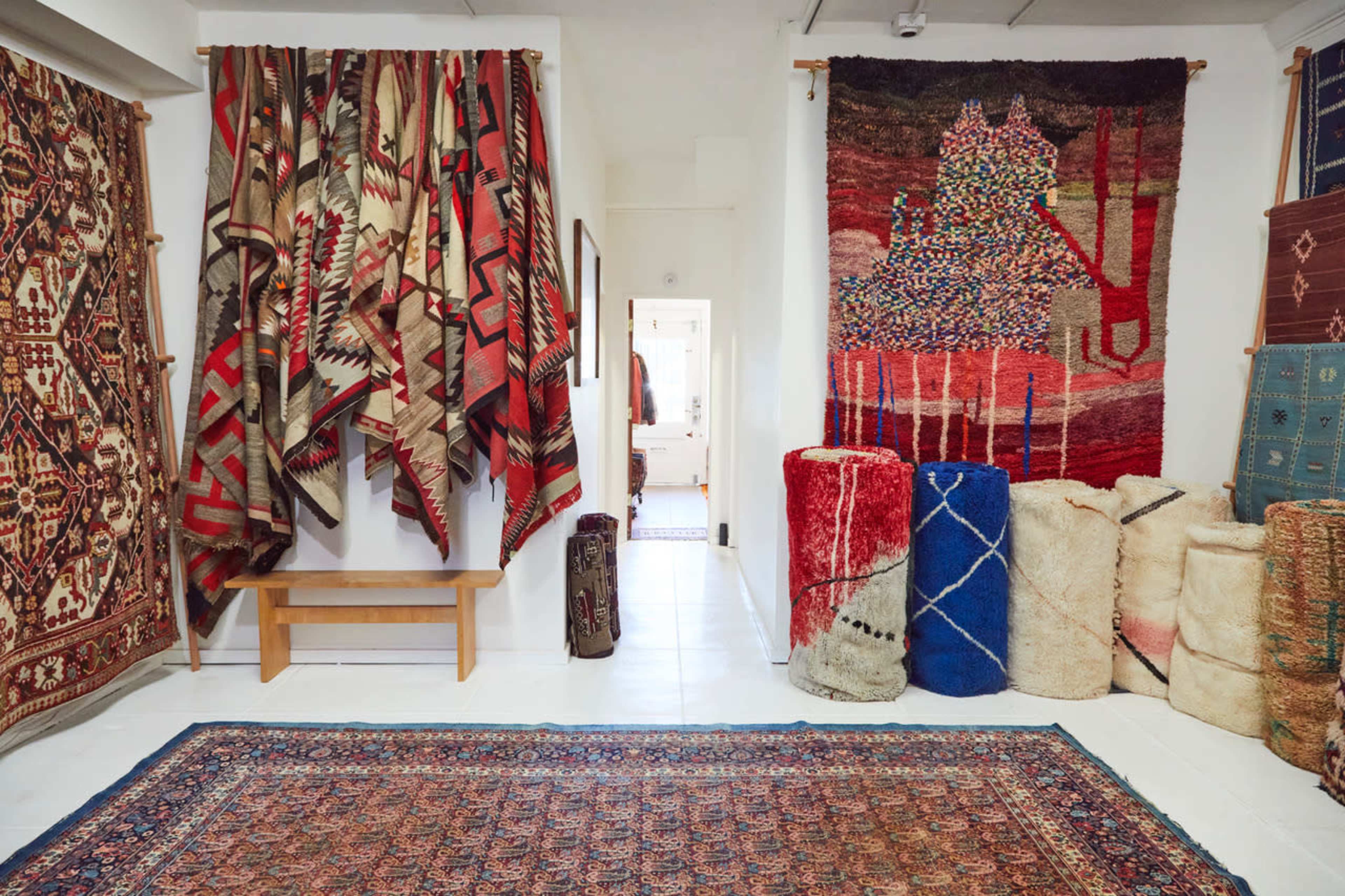 The image shows a room filled with various decorative textiles, including wall hangings and rolled rugs, arranged along the walls and on the floor.