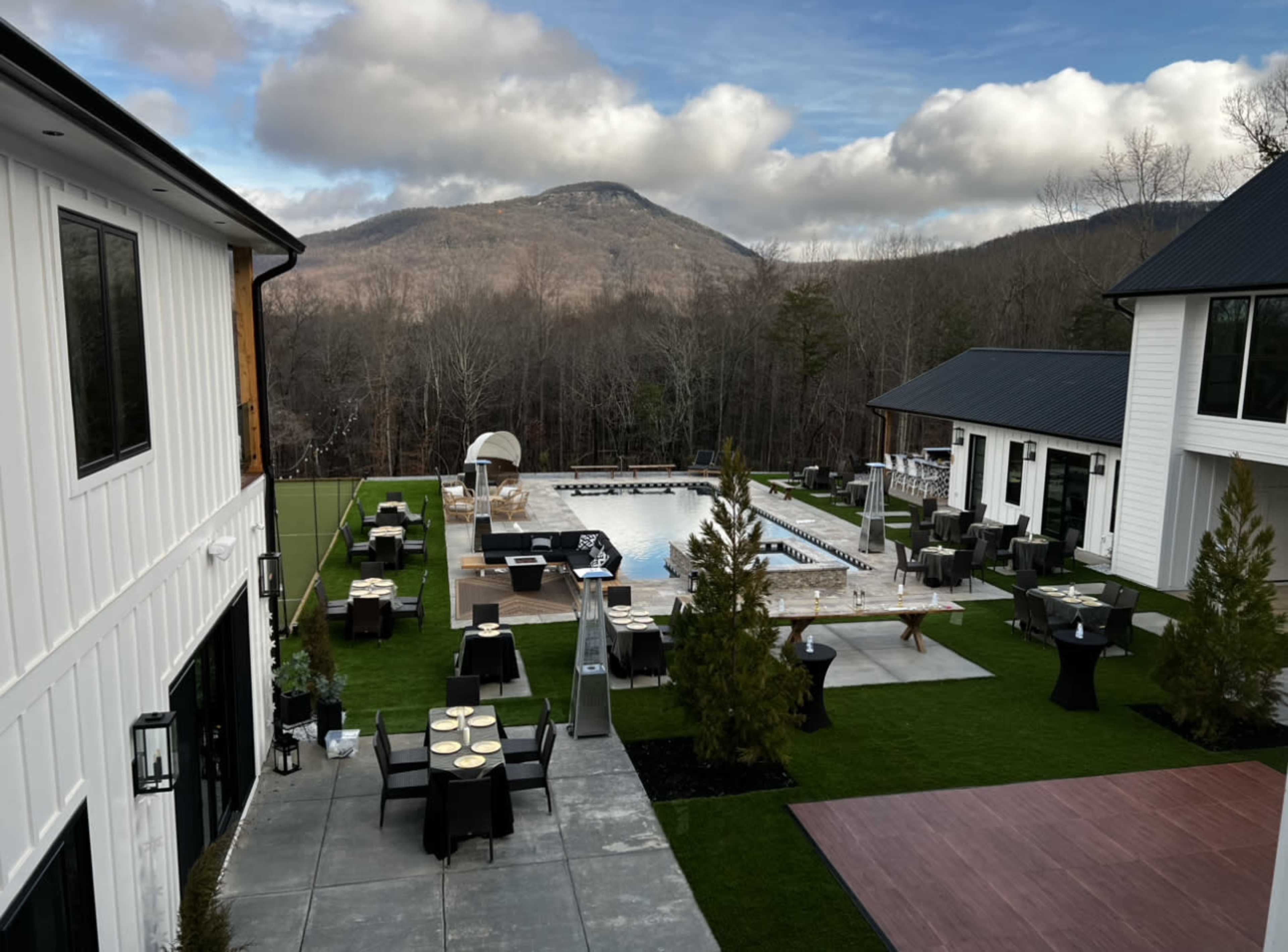 A modern outdoor area featuring a large pool, lounge furniture, and tables, surrounded by greenery and mountains in the background.