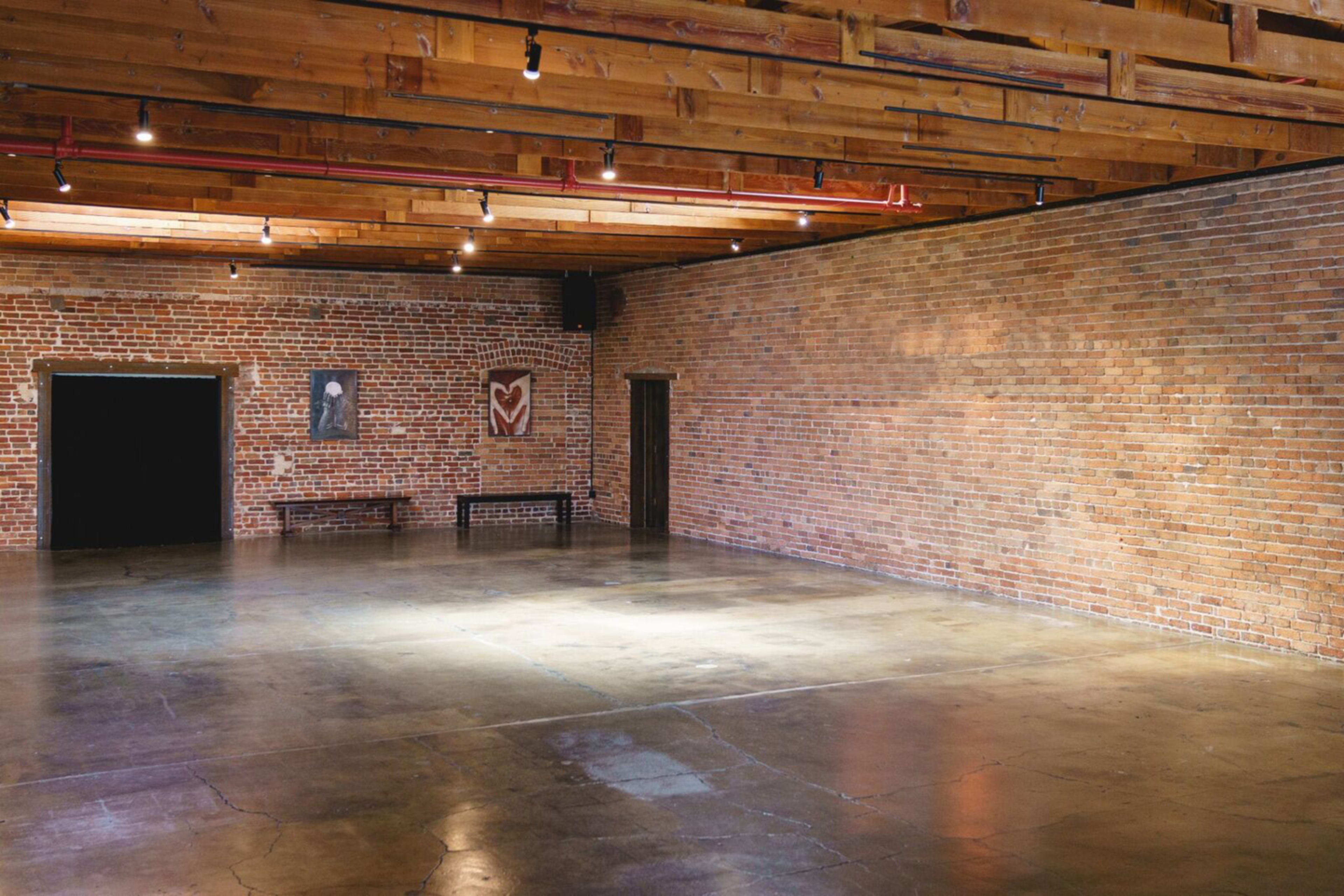 An empty room with brick walls, wooden beams, and polished concrete flooring, featuring a black doorway and a bench along one wall.