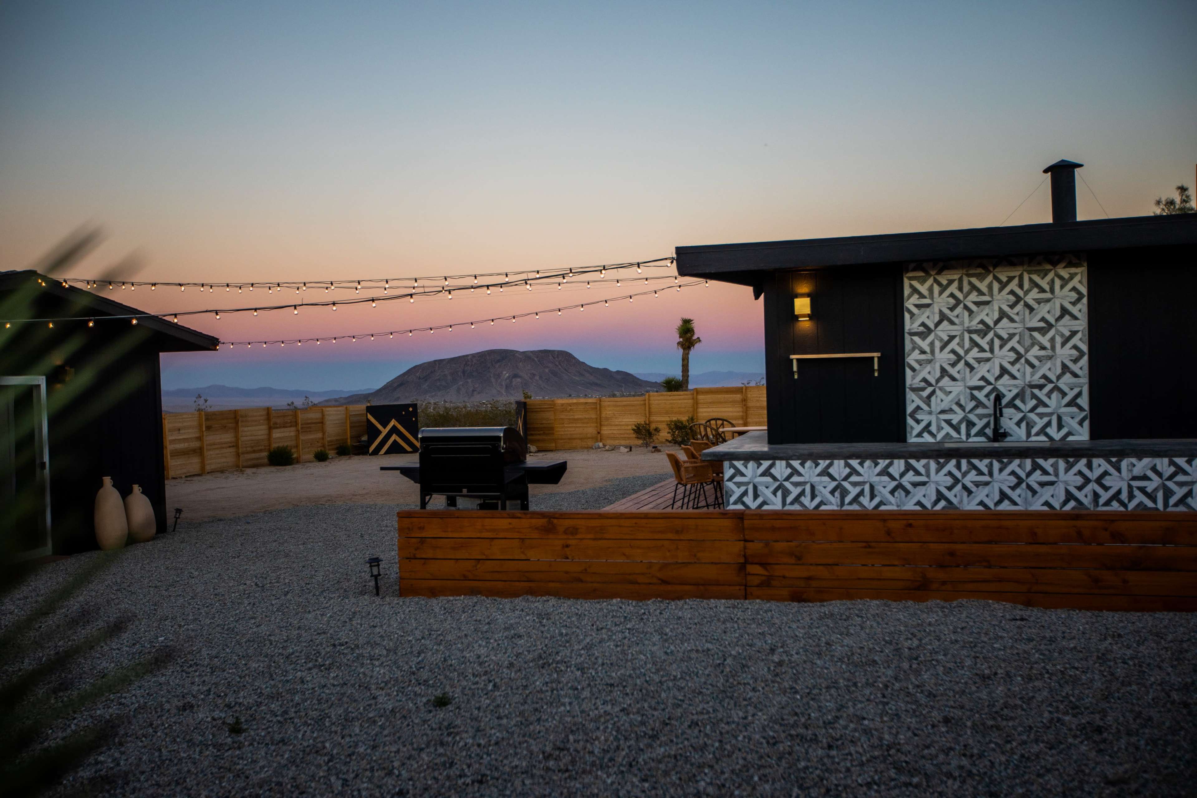 A modern home with patterned siding and a grill is situated in a gravel yard, framed by string lights and a mountain in the background at dusk.