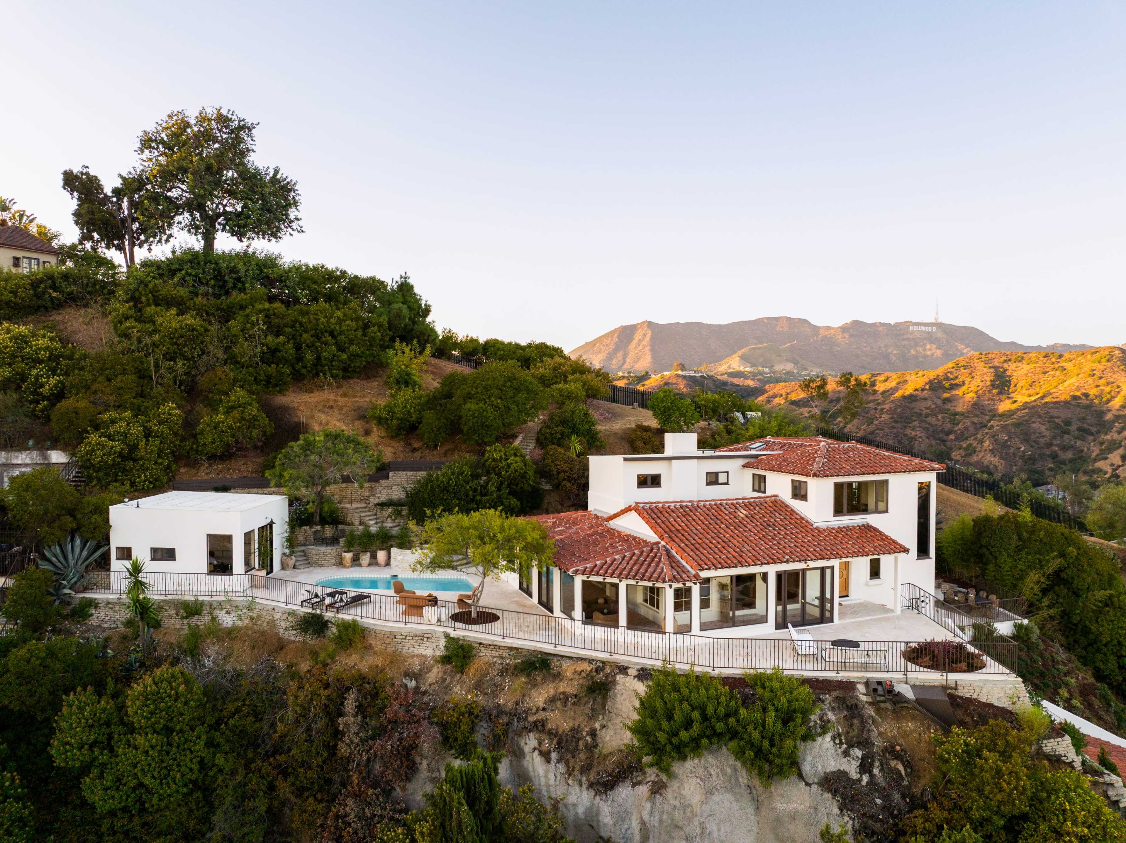 The image shows a modern white house with a red tile roof, surrounded by lush greenery and a pool, set against a mountainous backdrop.
