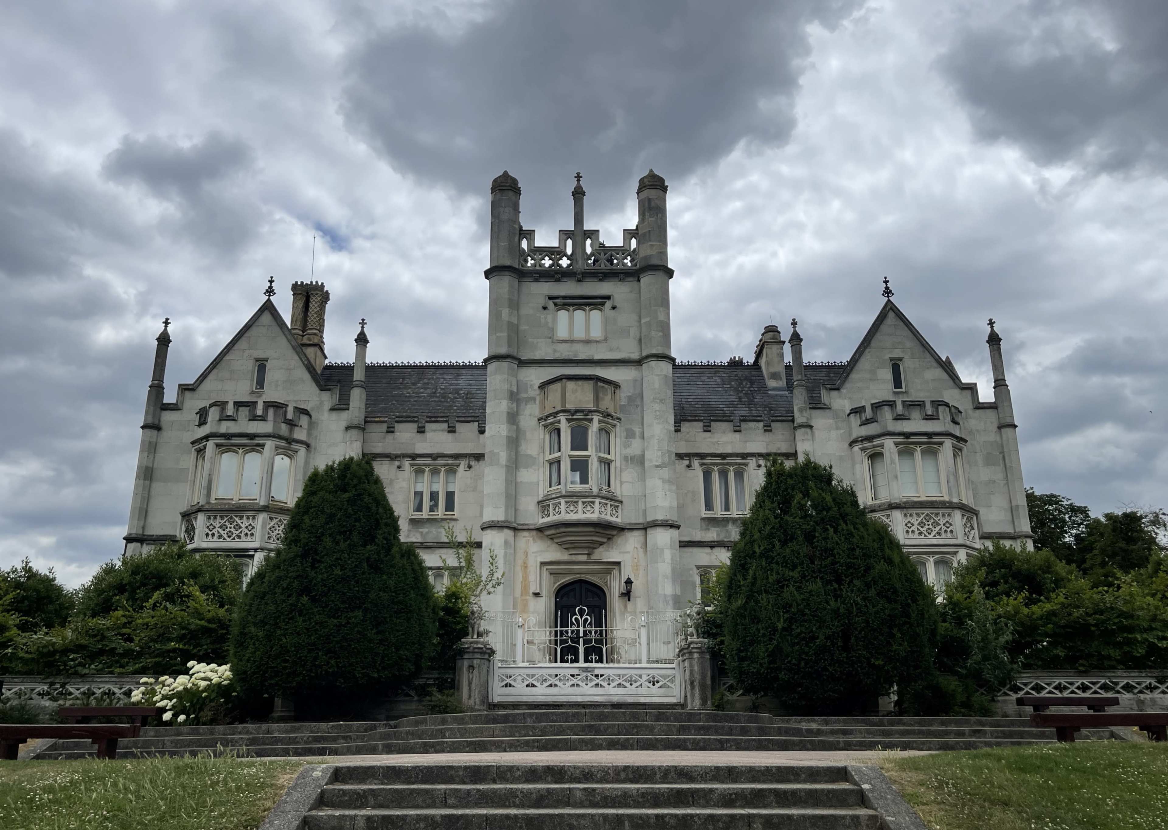 A large, ornate mansion stands before a staircase, framed by two tall hedges and set against a cloudy sky.