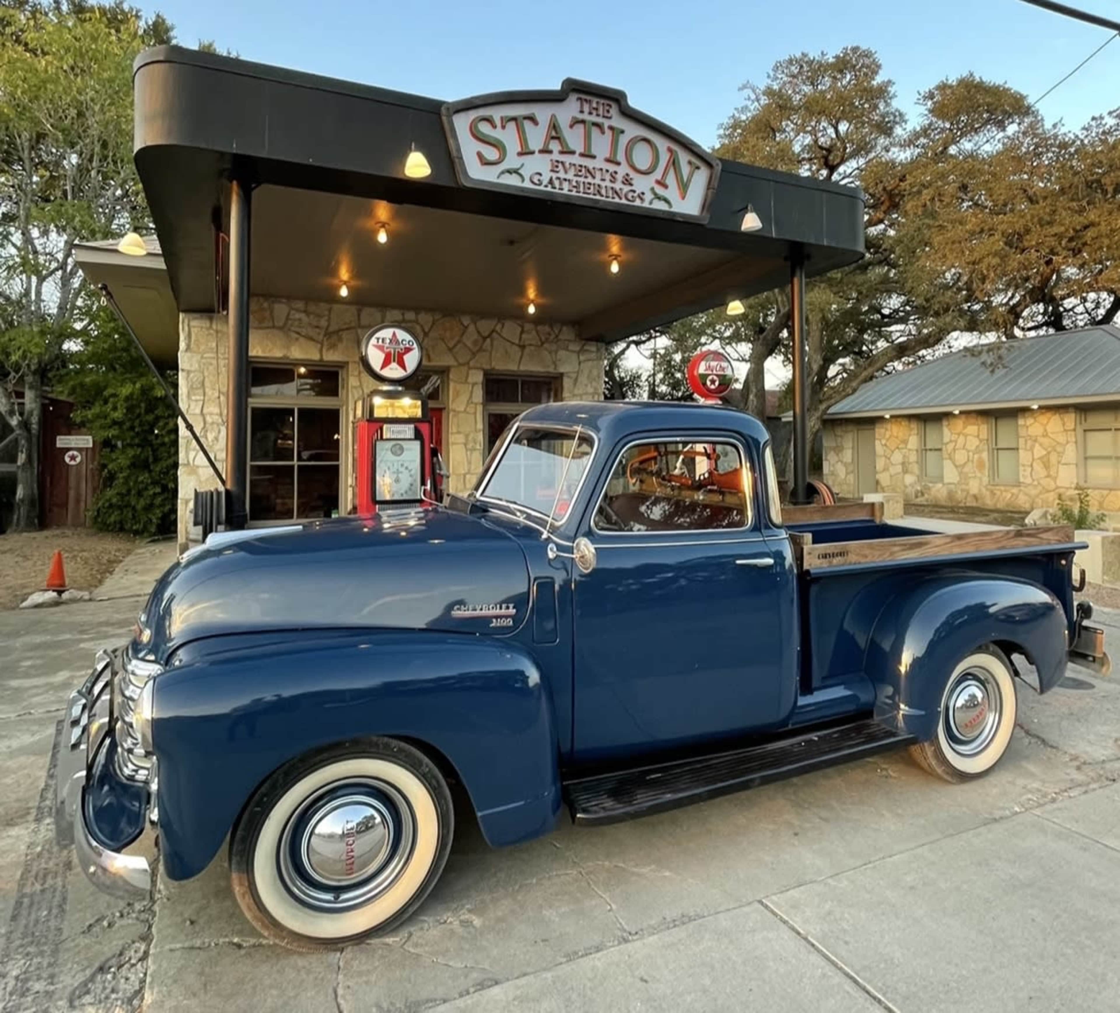 An old blue pickup truck is parked in front of a vintage gas station building with a stone exterior and a sign reading "The Station."