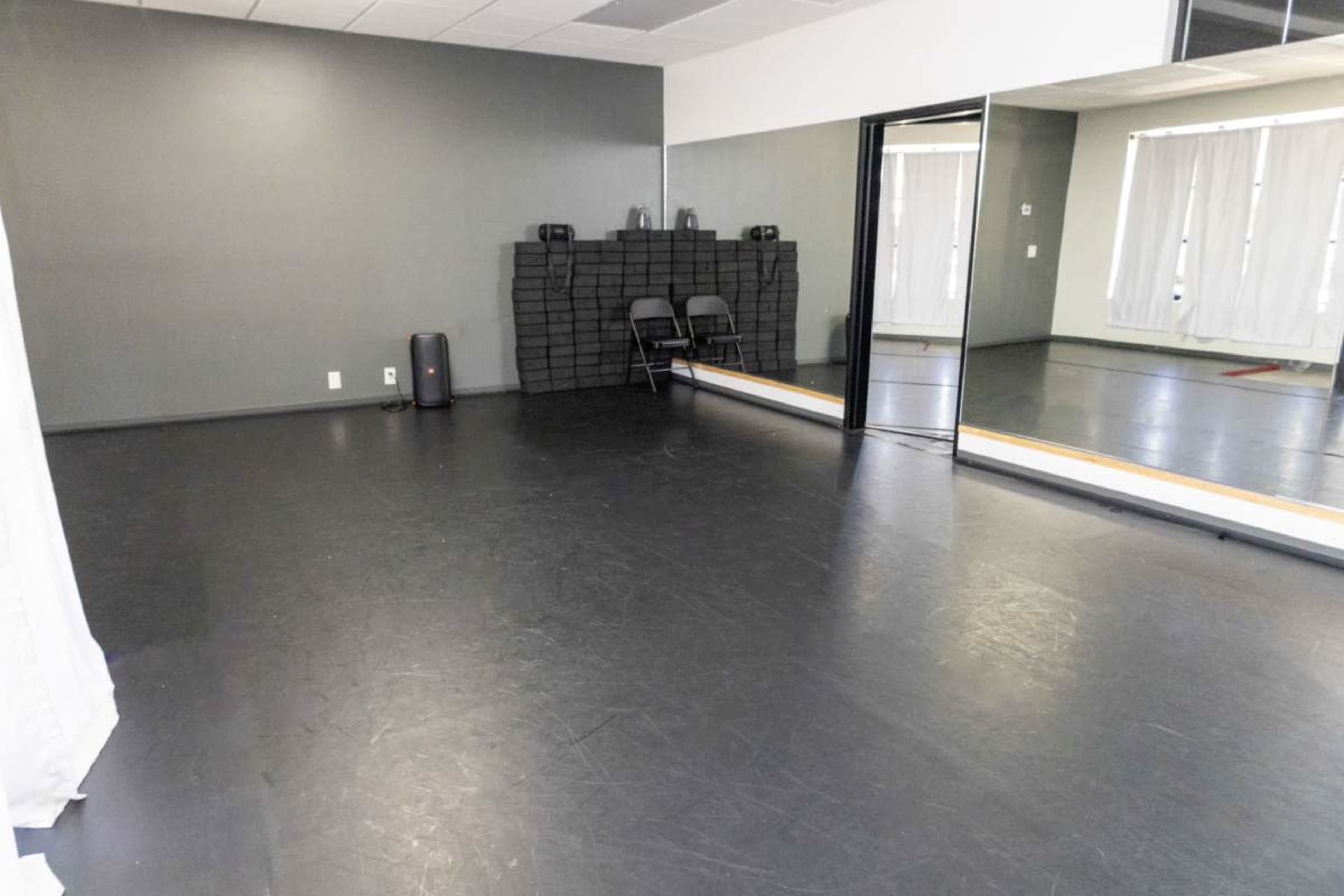 The image shows an empty dance studio with black flooring, a large mirror, and stacks of mats against a gray wall.