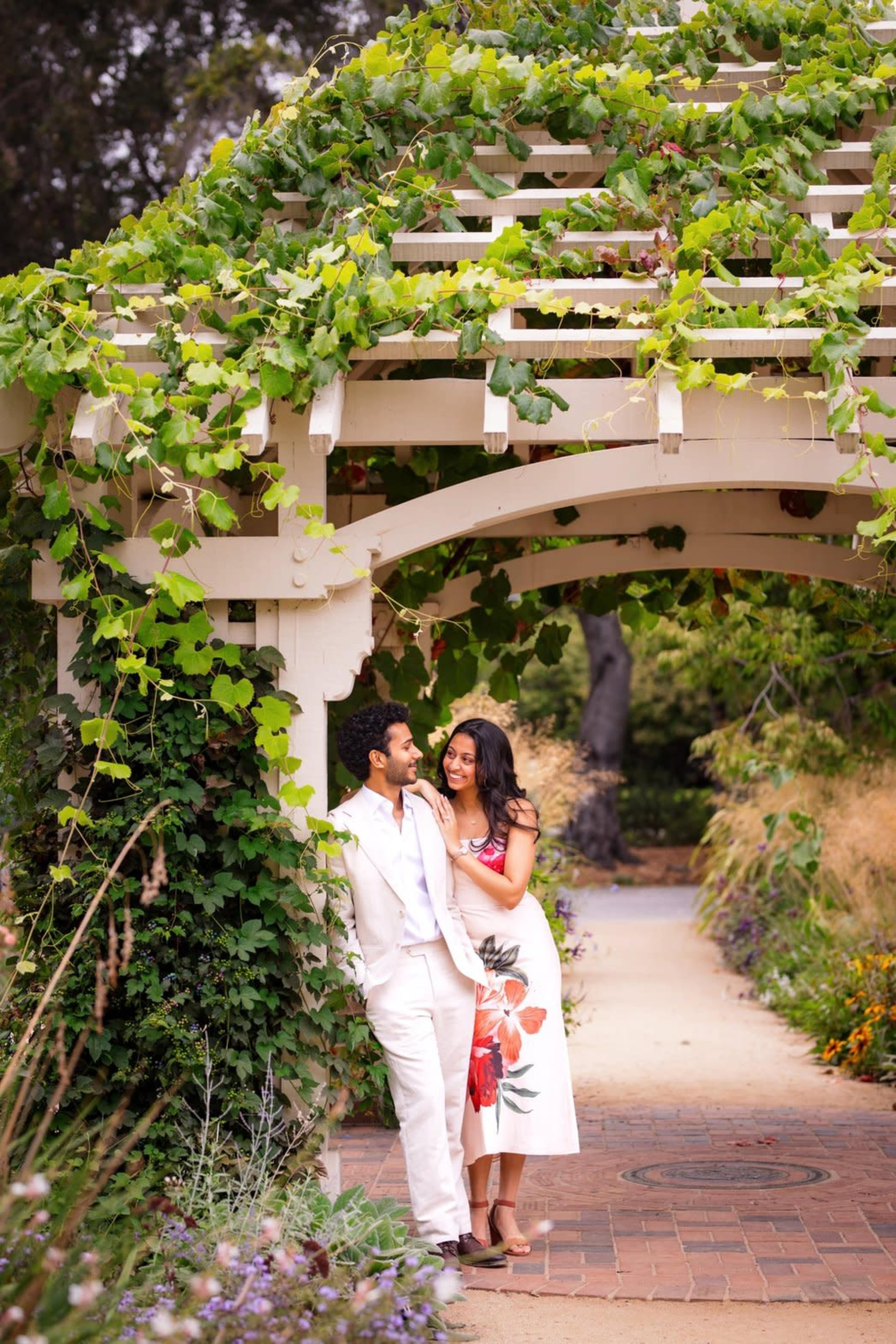 A couple stands closely under a vine-covered arbor in a garden setting.