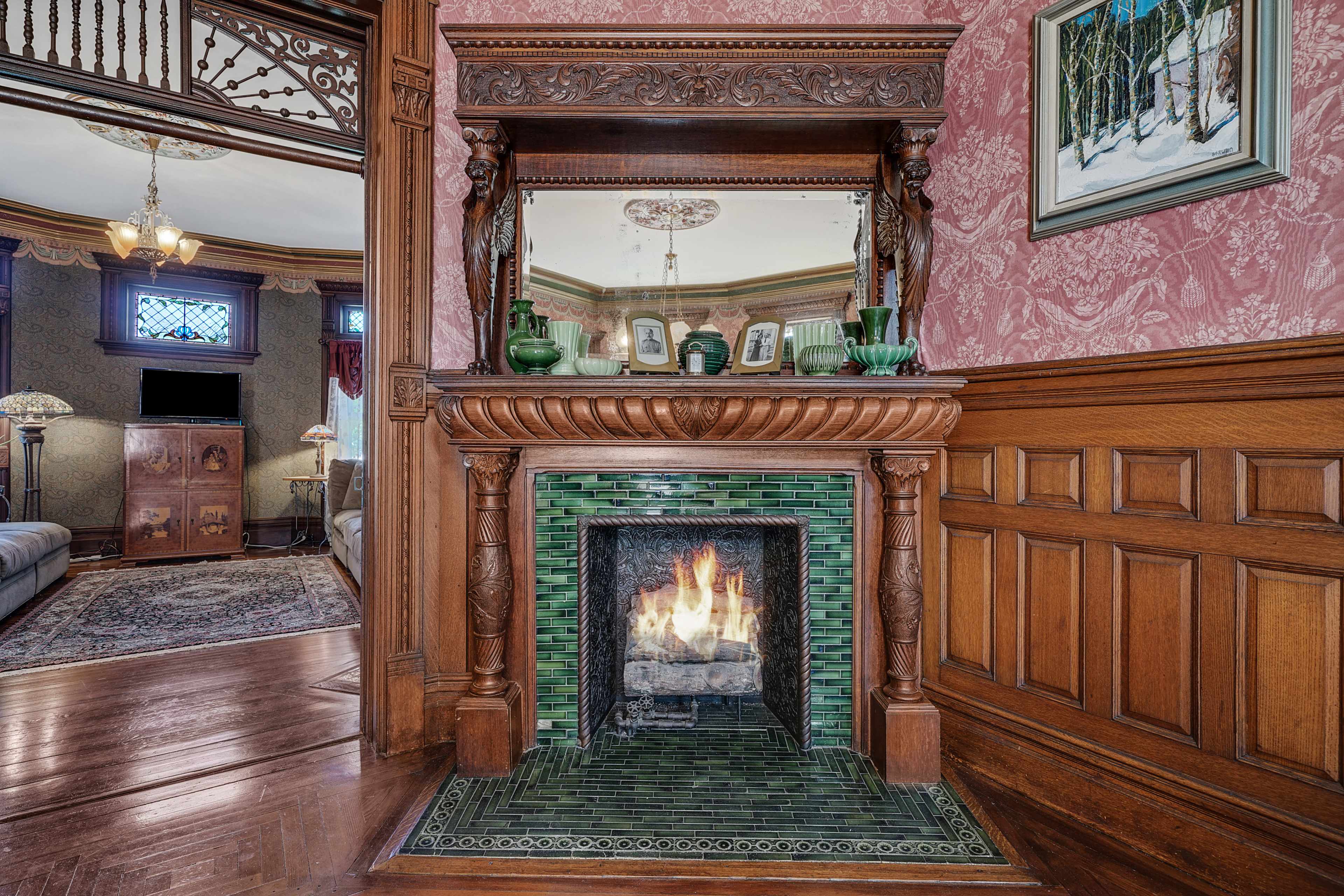 An ornate wooden fireplace mantel with intricate carvings and green ceramic tiles, featuring a lit fire, situated in a richly adorned room.