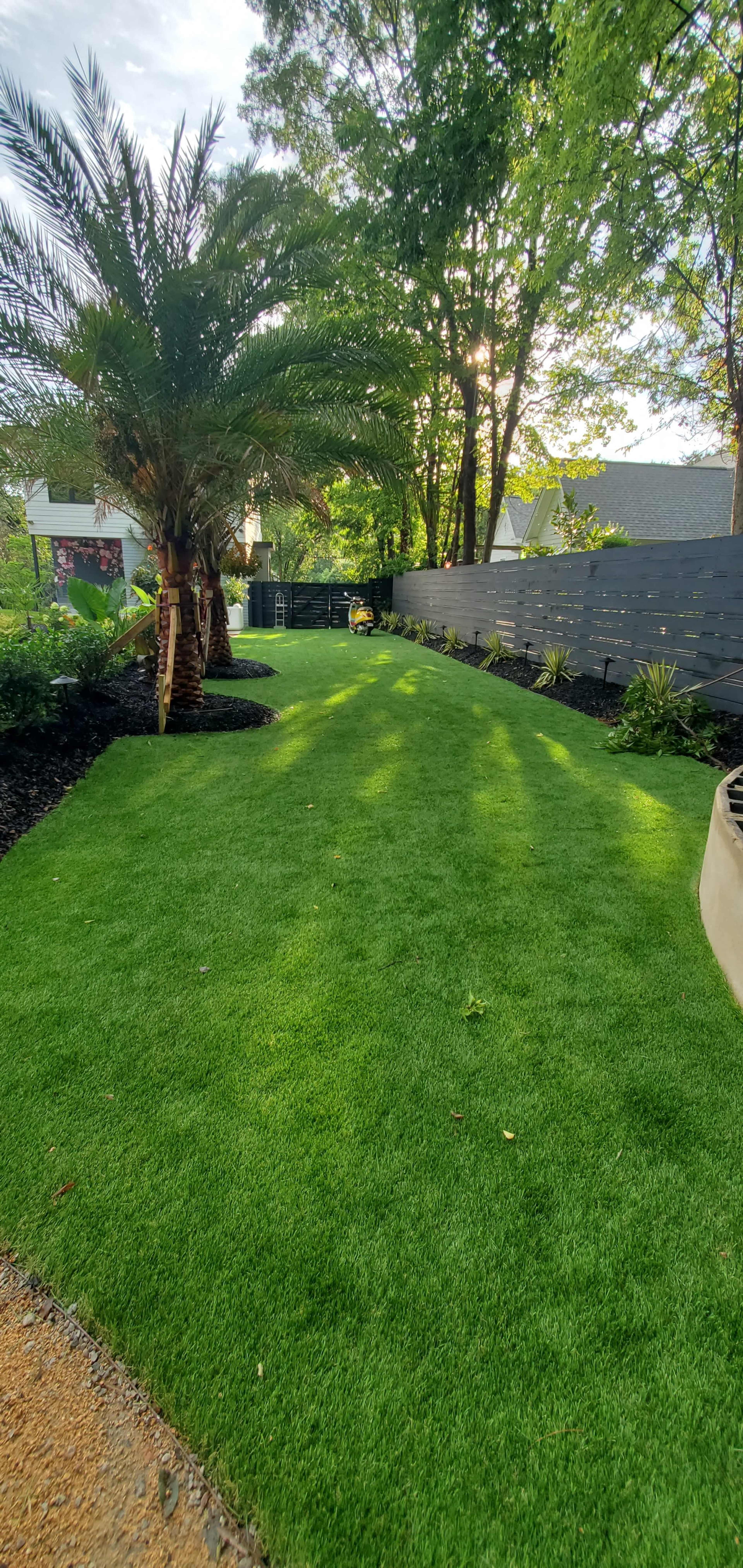 The image shows a well-manicured lawn with artificial grass, bordered by a palm tree and various plants, against a backdrop of a garden wall and trees.