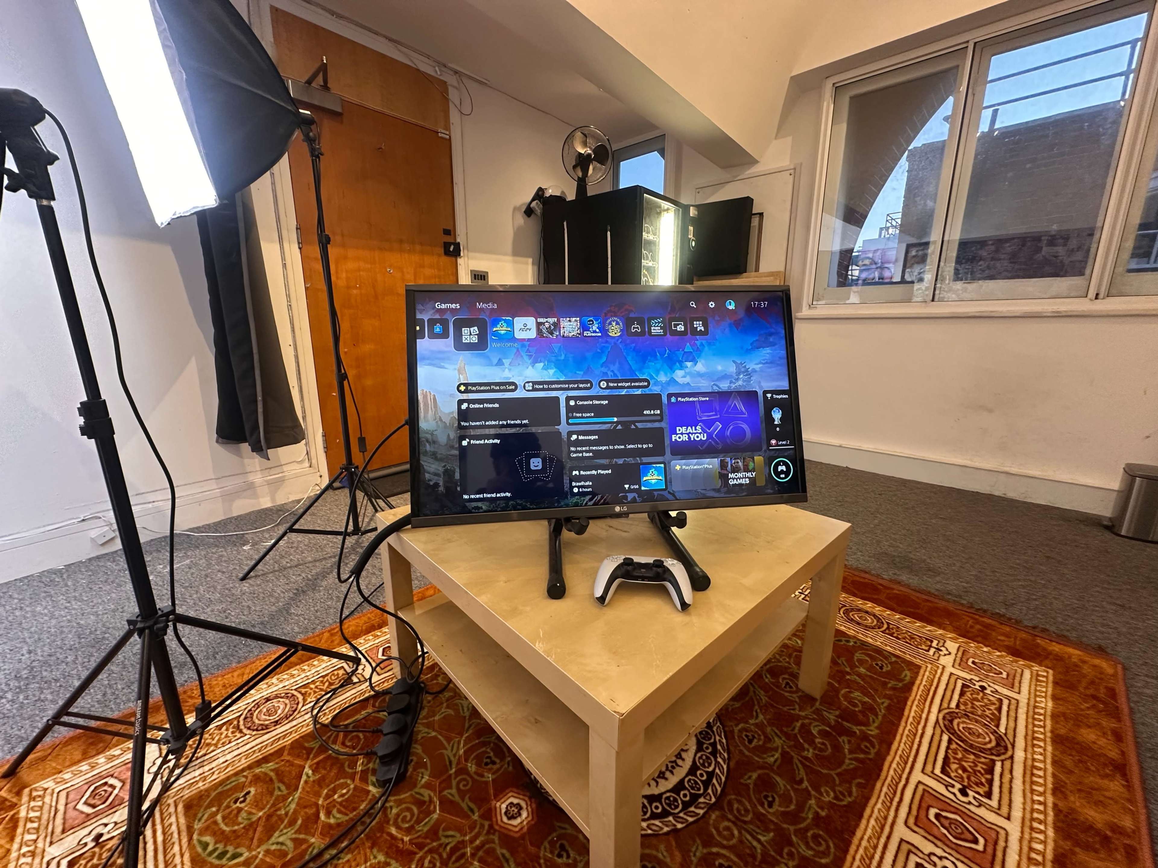 A gaming monitor displays a user interface on a small table in a well-lit room with a window and various studio lights set up.