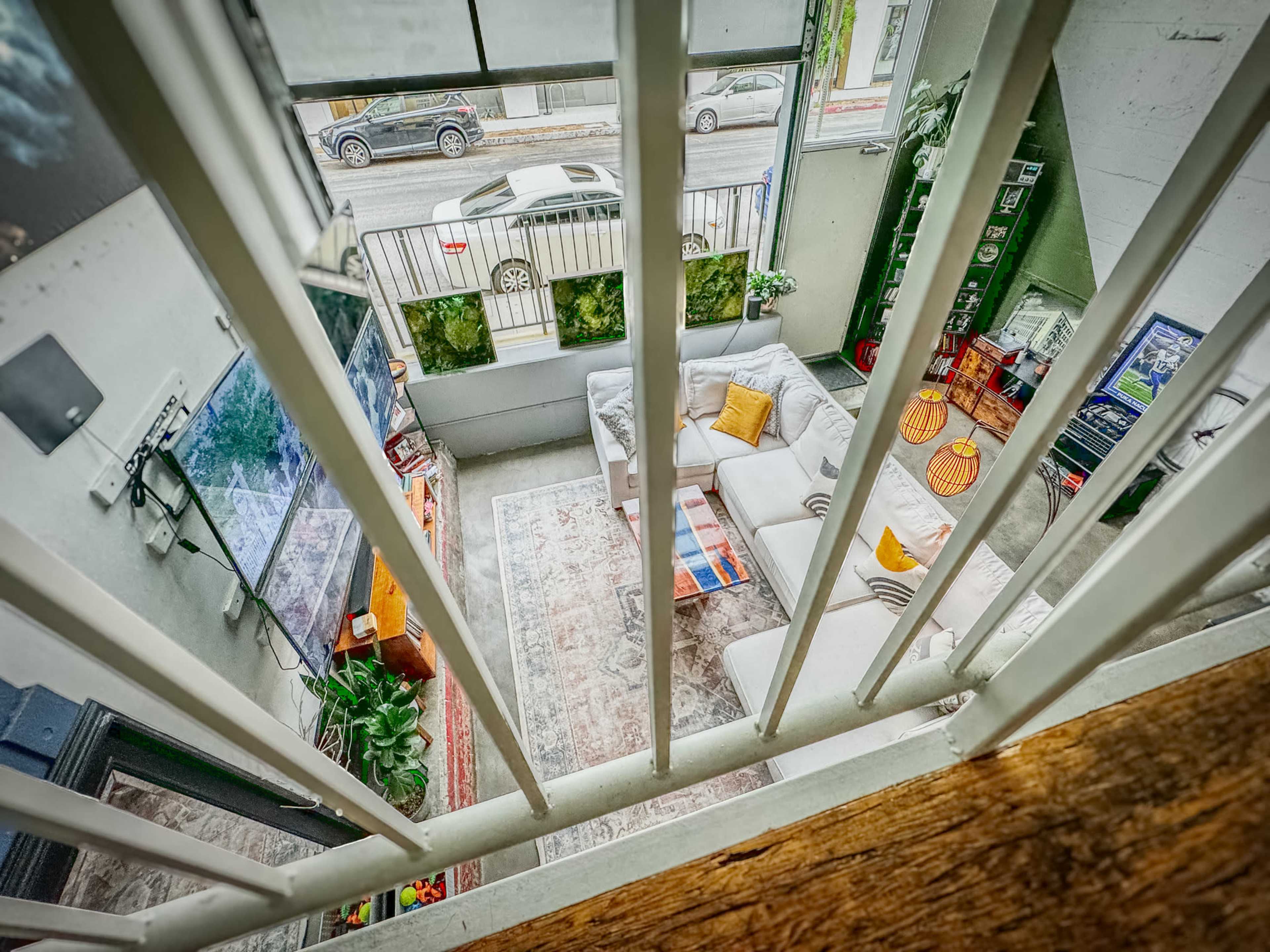 The image shows a living room viewed from above, featuring a sectional sofa, a television, and various plants arranged on a patterned rug.