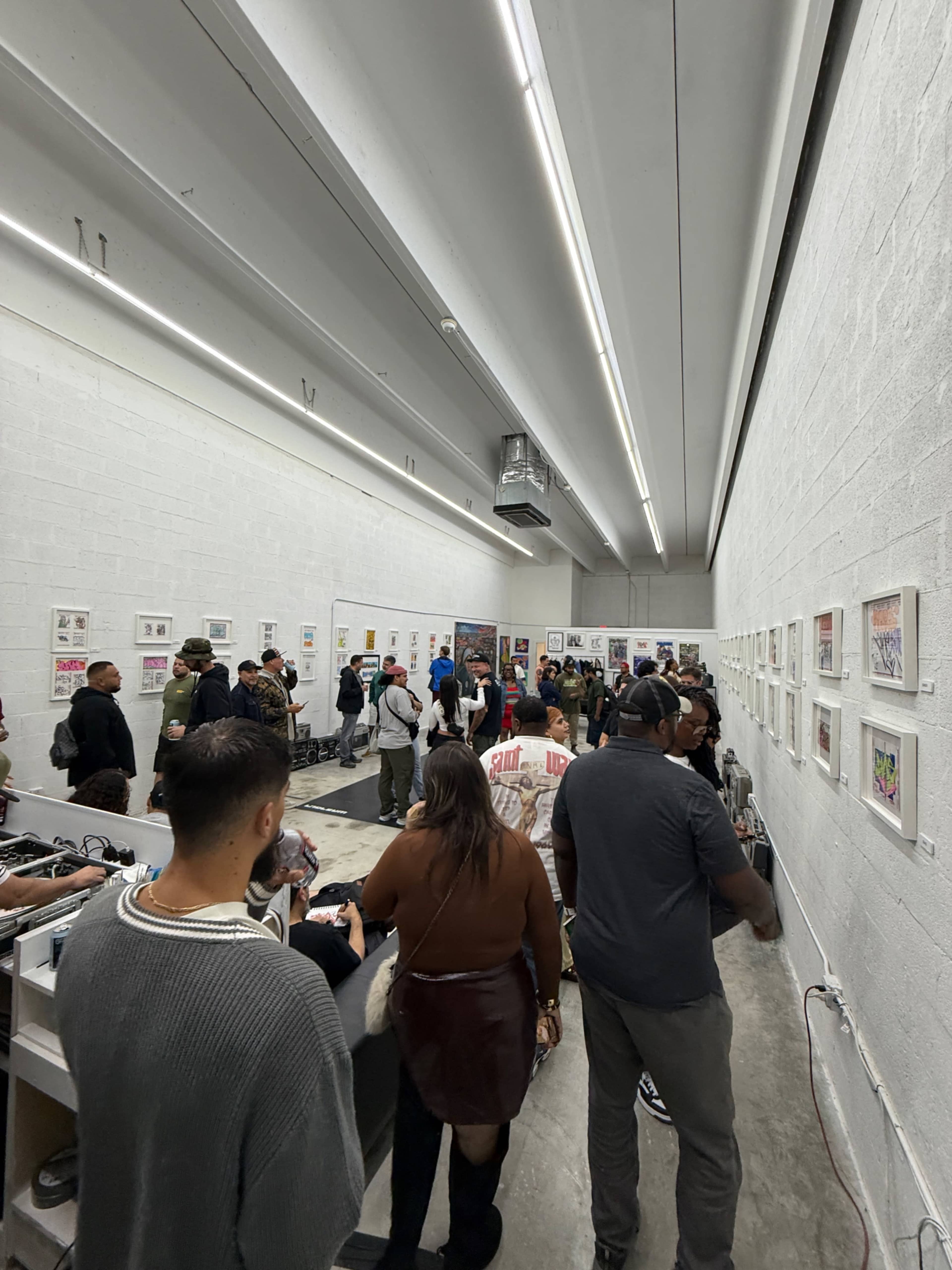 A group of people is gathered in an art gallery, examining framed artwork displayed on the white walls.