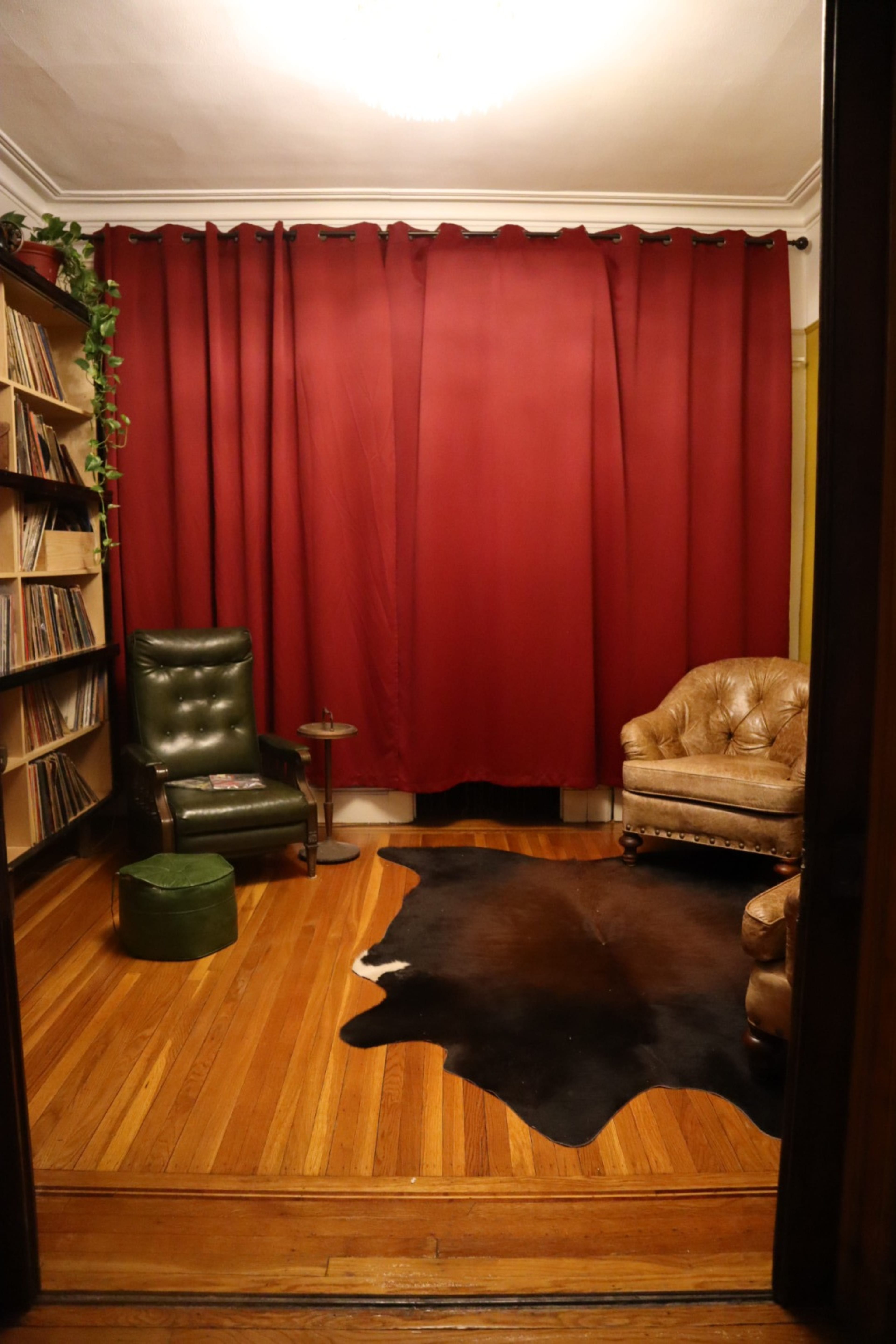 The image shows a room with a wooden floor, a bookshelf filled with records, a green chair, a brown leather armchair, a small side table, and red curtains.