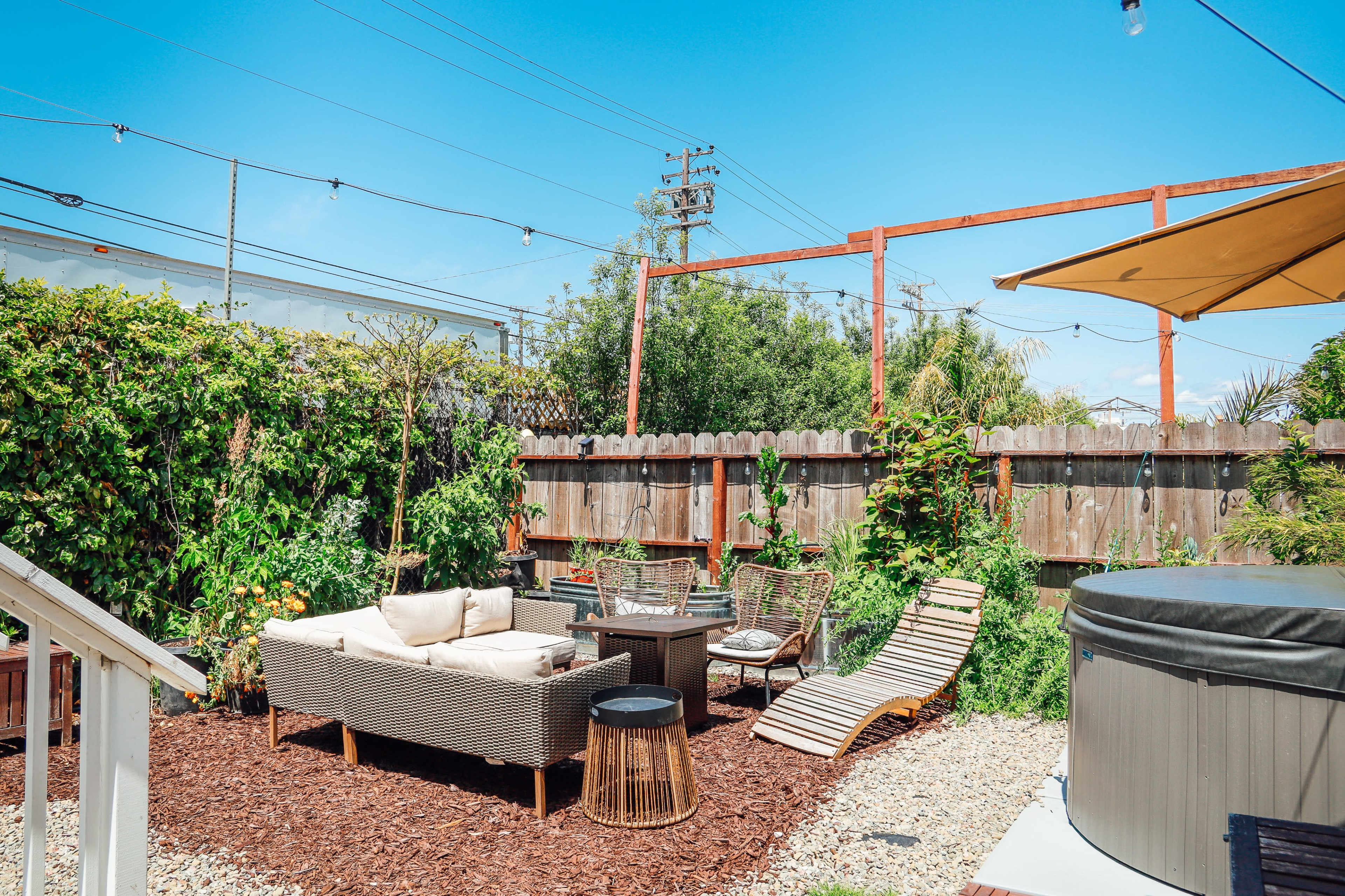 A landscaped outdoor space features a seating area with a sofa, chairs, and a lounge chair surrounded by greenery and a hot tub.
