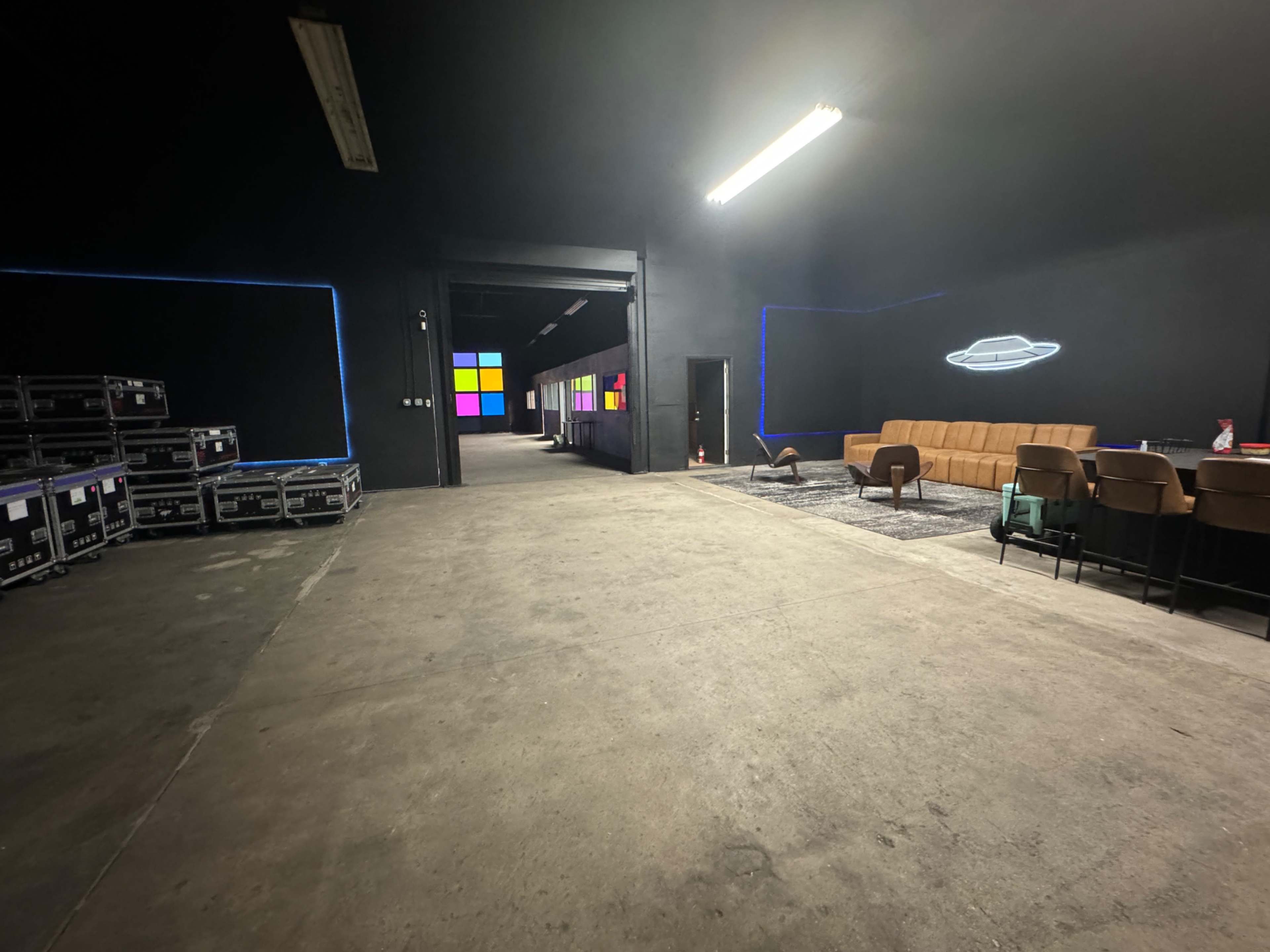 The image depicts an industrial space featuring a seating area with brown chairs and a couch, surrounded by illuminated walls and equipment cases stacked along the left side.