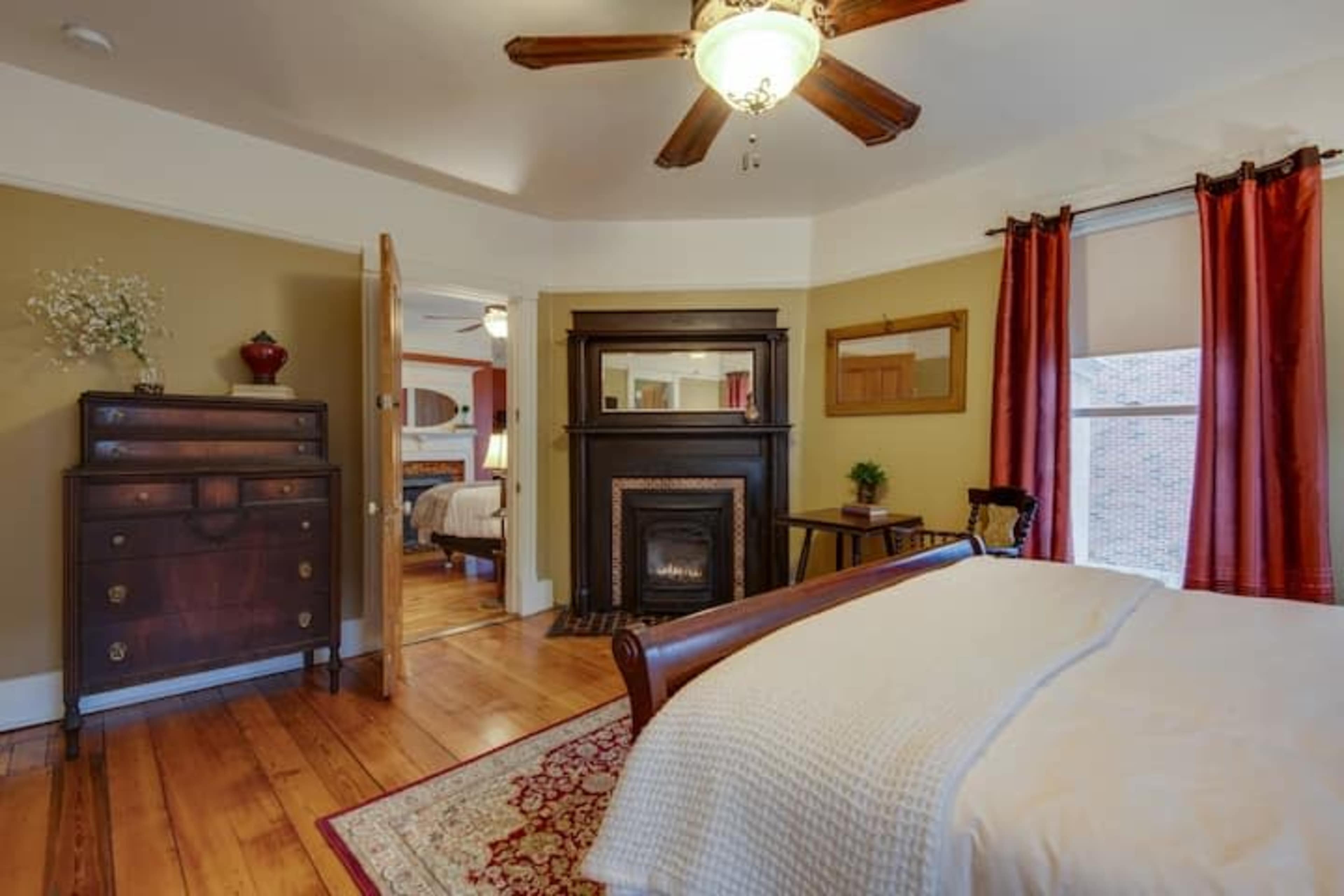 A well-lit bedroom featuring a wooden bed, a fireplace with a decorative mantel, and red curtains.