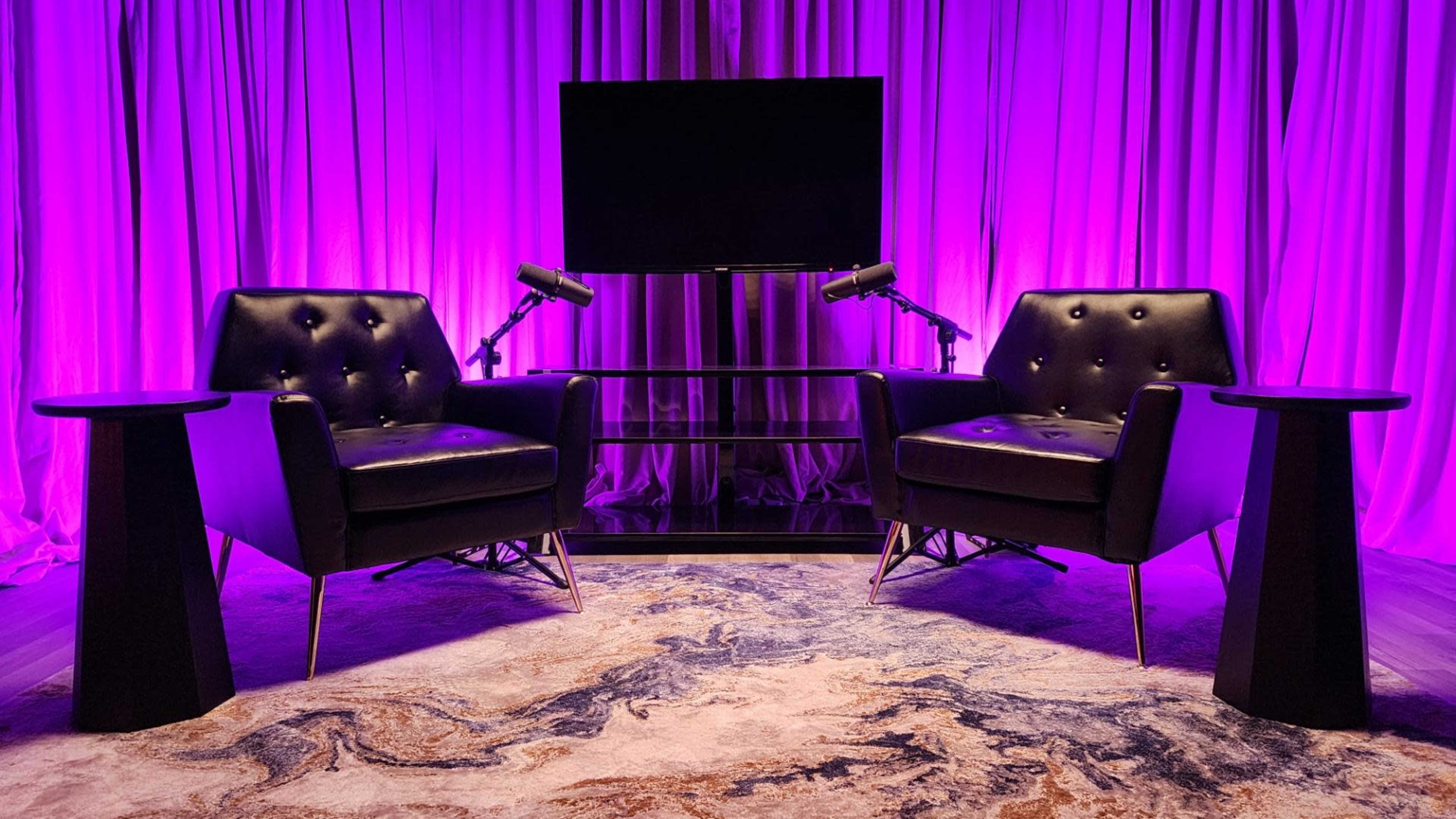 The image features two black armchairs positioned facing each other, with microphones on stands and a large screen in the background, all set against purple drapery.