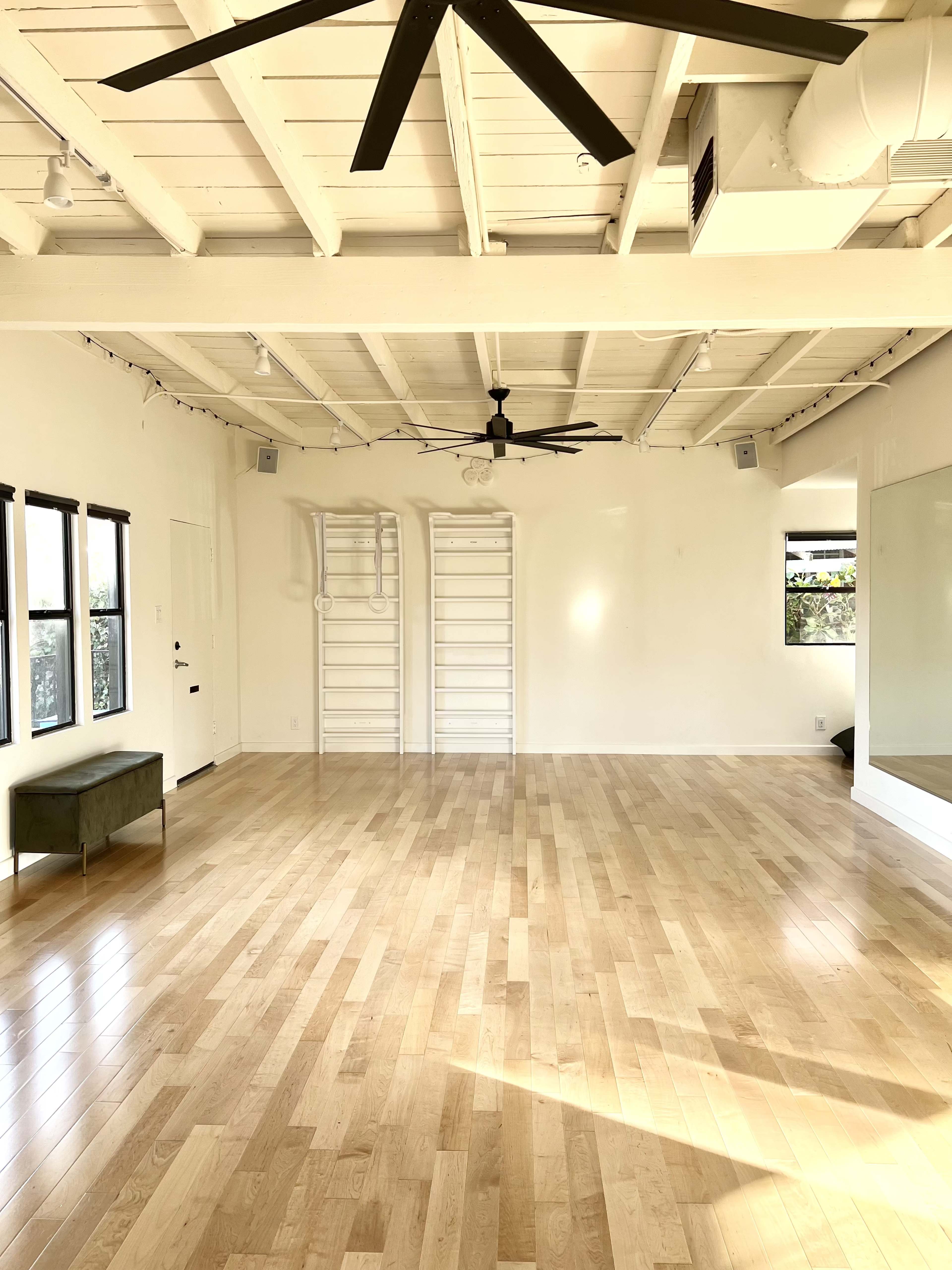 A spacious, empty room with wooden floors, white walls, and a mirrored wall, featuring a ceiling fan and a green bench.