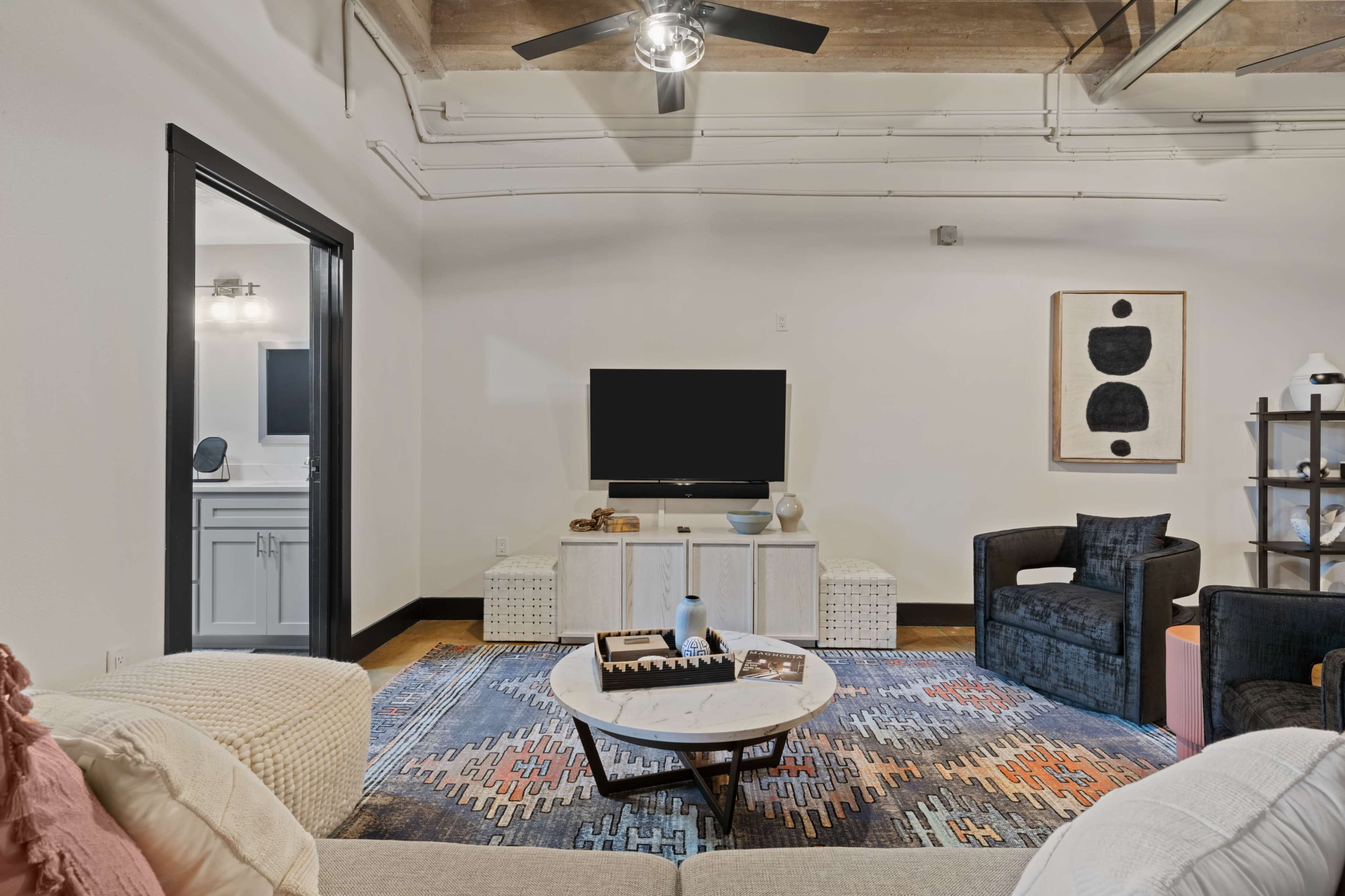 A modern living room with a flat-screen TV mounted on the wall, a neutral-colored sofa, a round coffee table, and two armchairs situated around a patterned area rug.