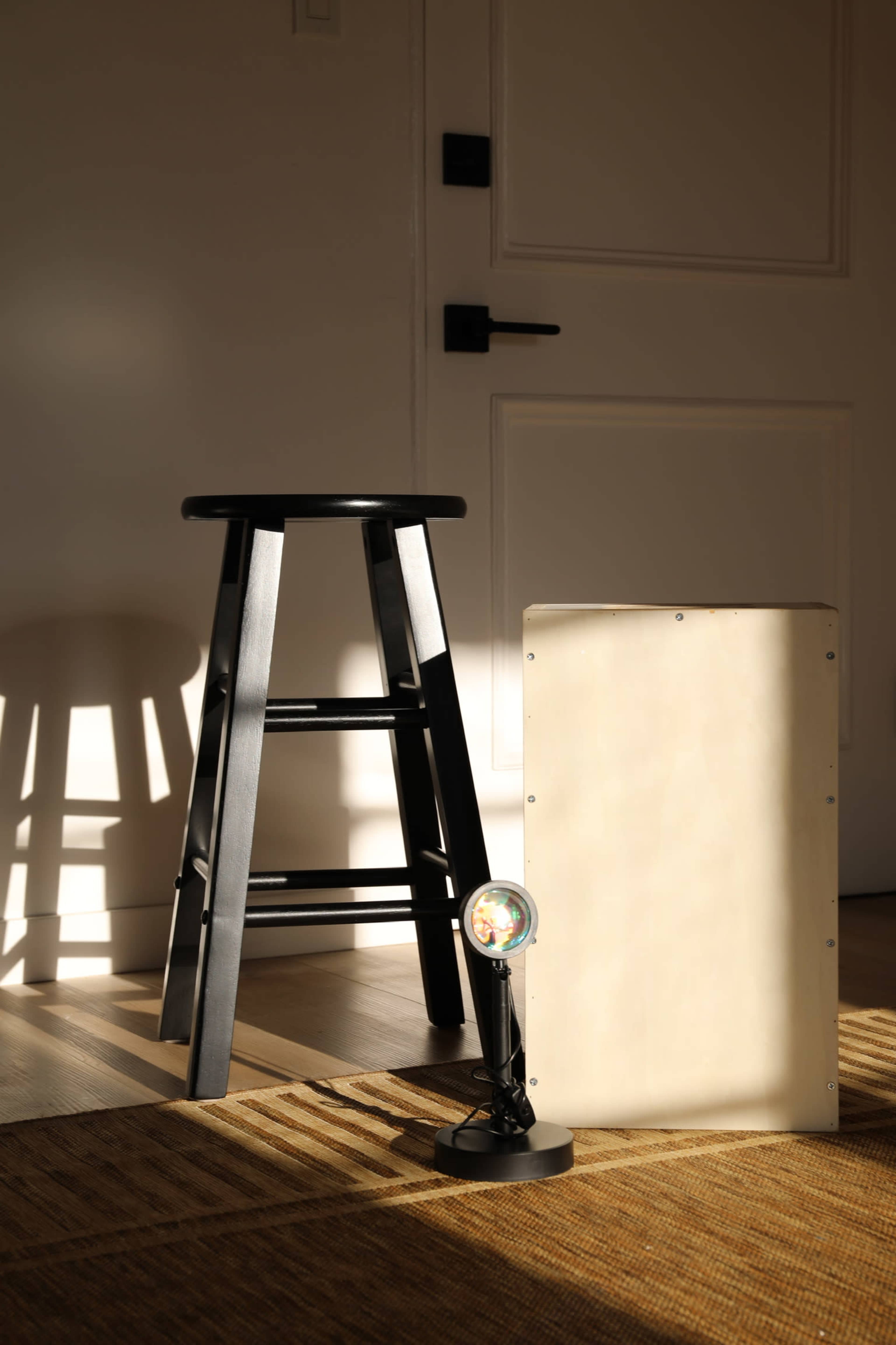 A black stool, a light fixture, and a plain panel are arranged on a wooden floor, illuminated by soft sunlight.