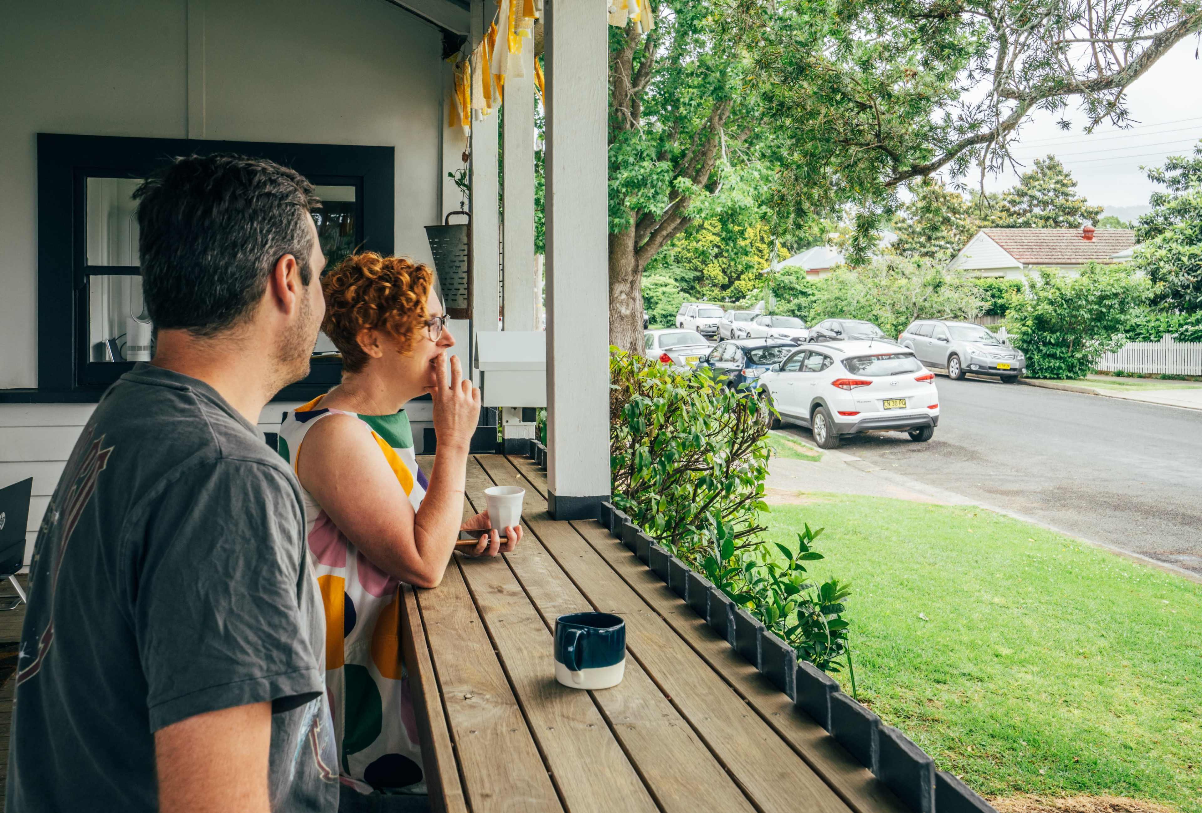 A man and a woman stand on a porch, sipping from cups and looking at the street outside.