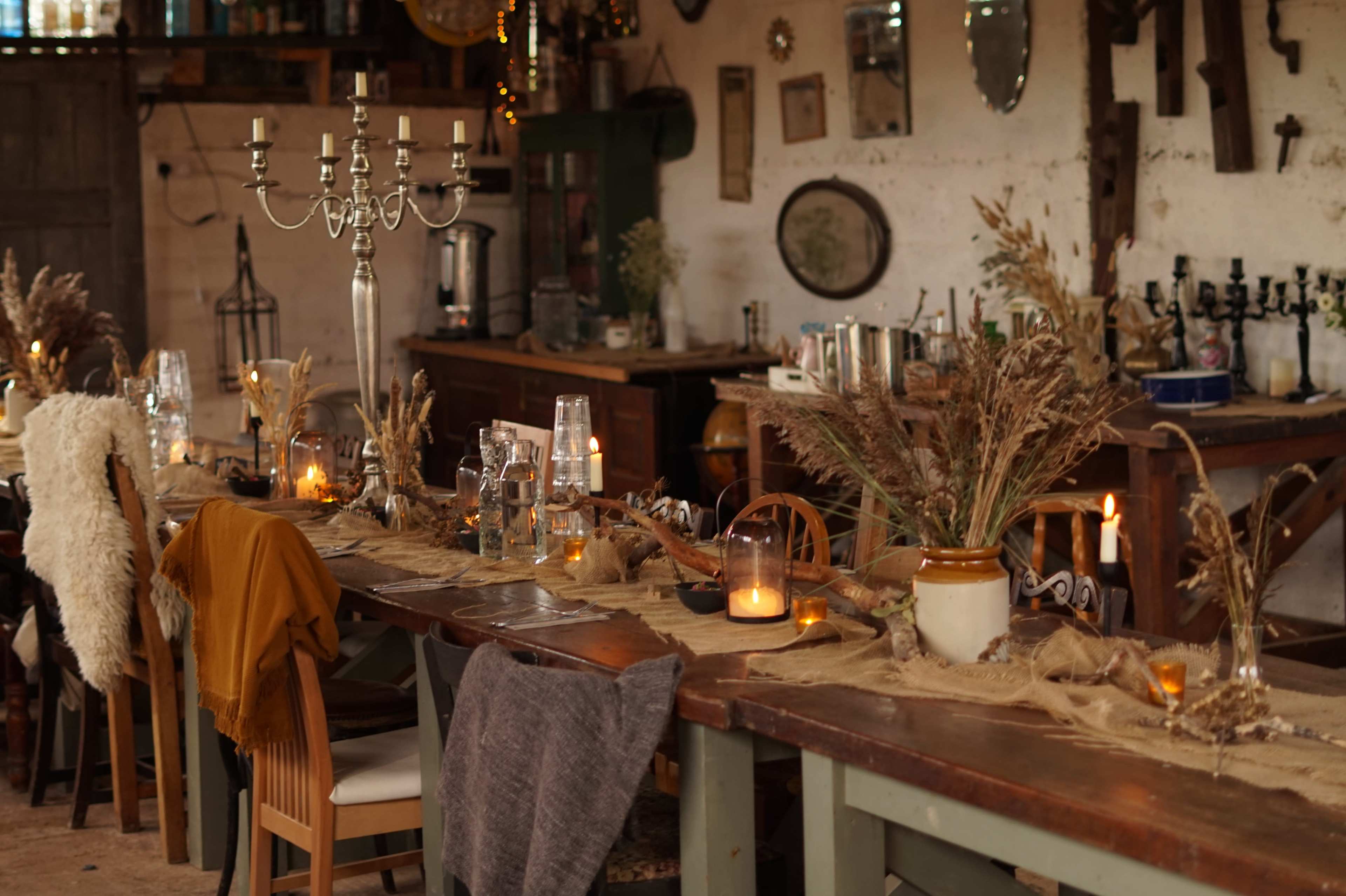 A rustic dining table is decorated with dried plants, candles, and glassware in a warmly lit room featuring vintage decor.