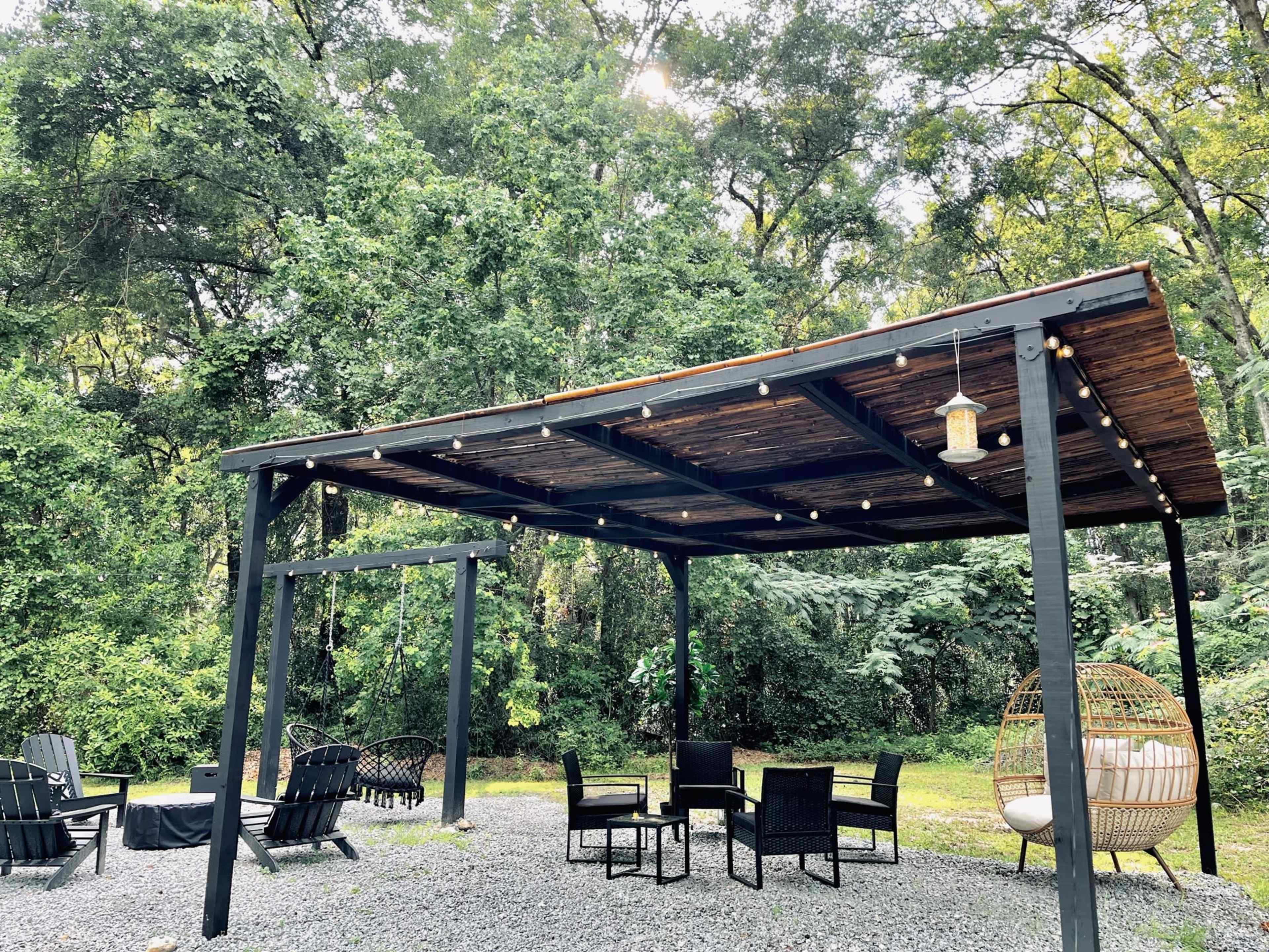 A shaded outdoor seating area features a wooden pergola, string lights, and a hanging chair surrounded by trees.