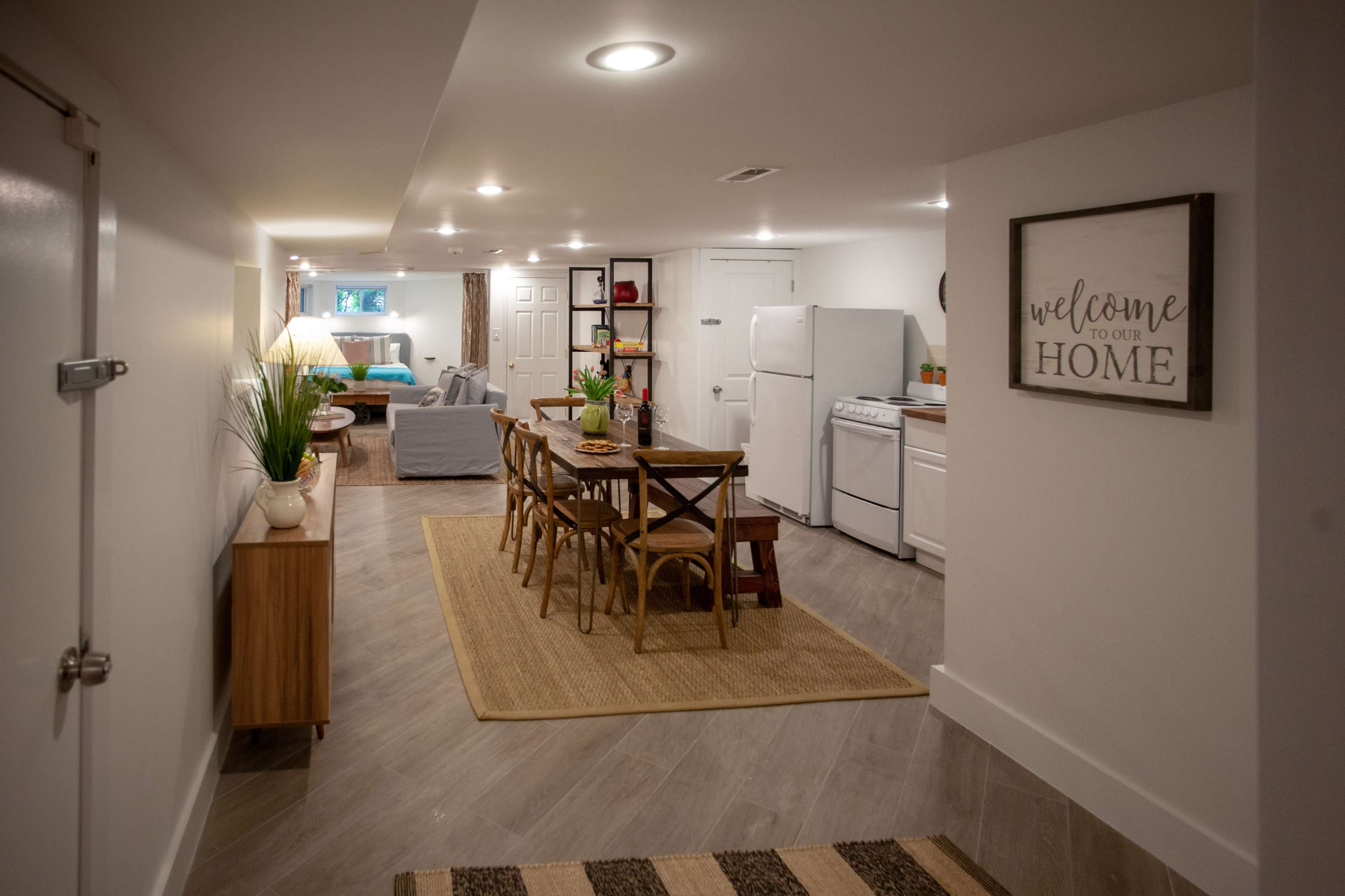 A well-lit basement area features a dining table with chairs, a kitchen with white appliances, and a cozy living space in the background.