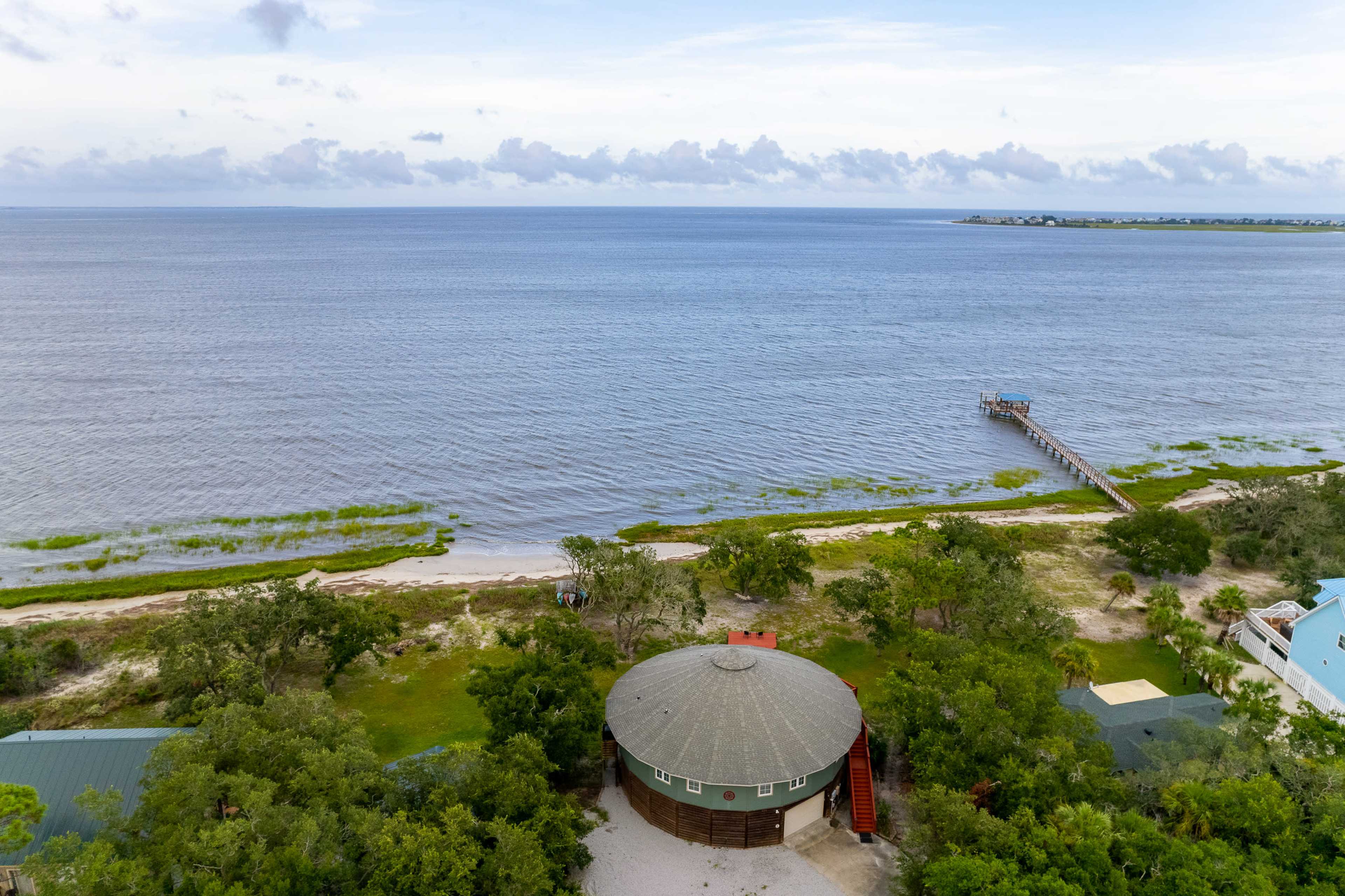 An aerial view shows a round structure near the shoreline with a dock extending into the water, surrounded by vegetation and a calm sea.