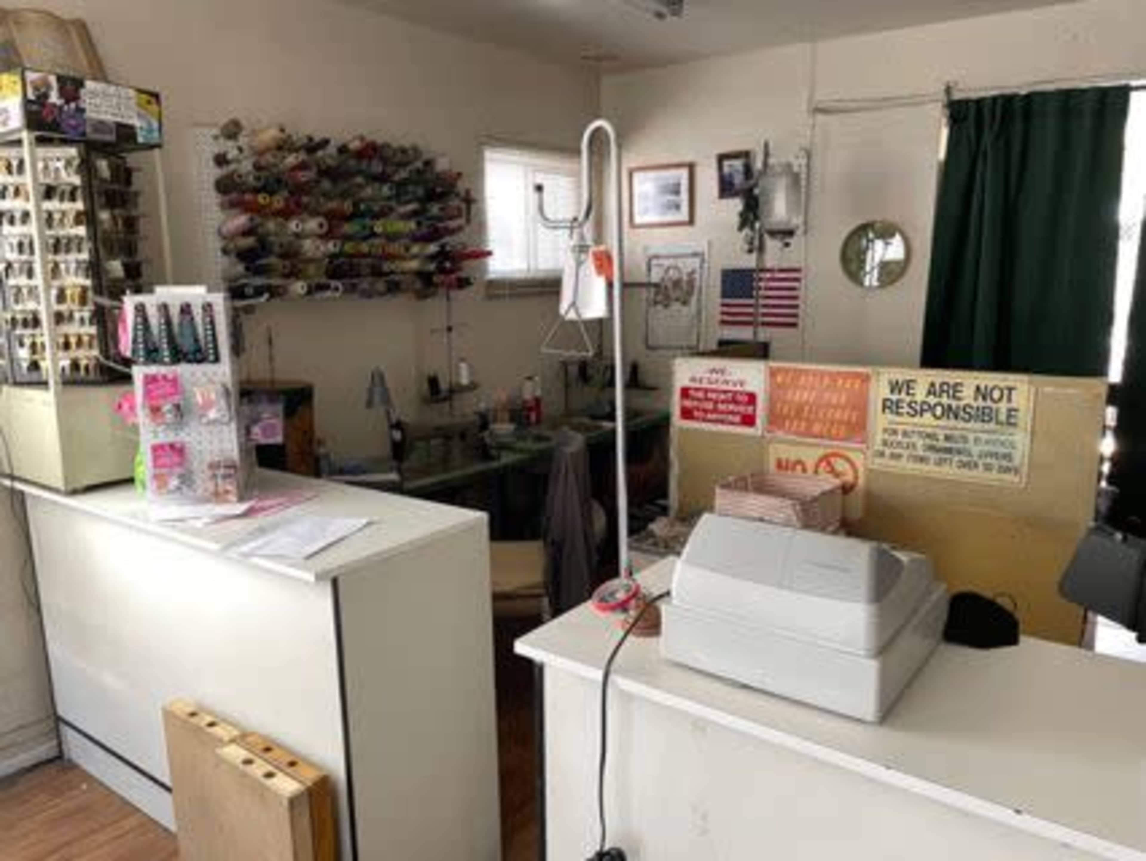The image shows the interior of a small shop with a counter, a sewing area in the background, and various items displayed on the walls and shelves.
