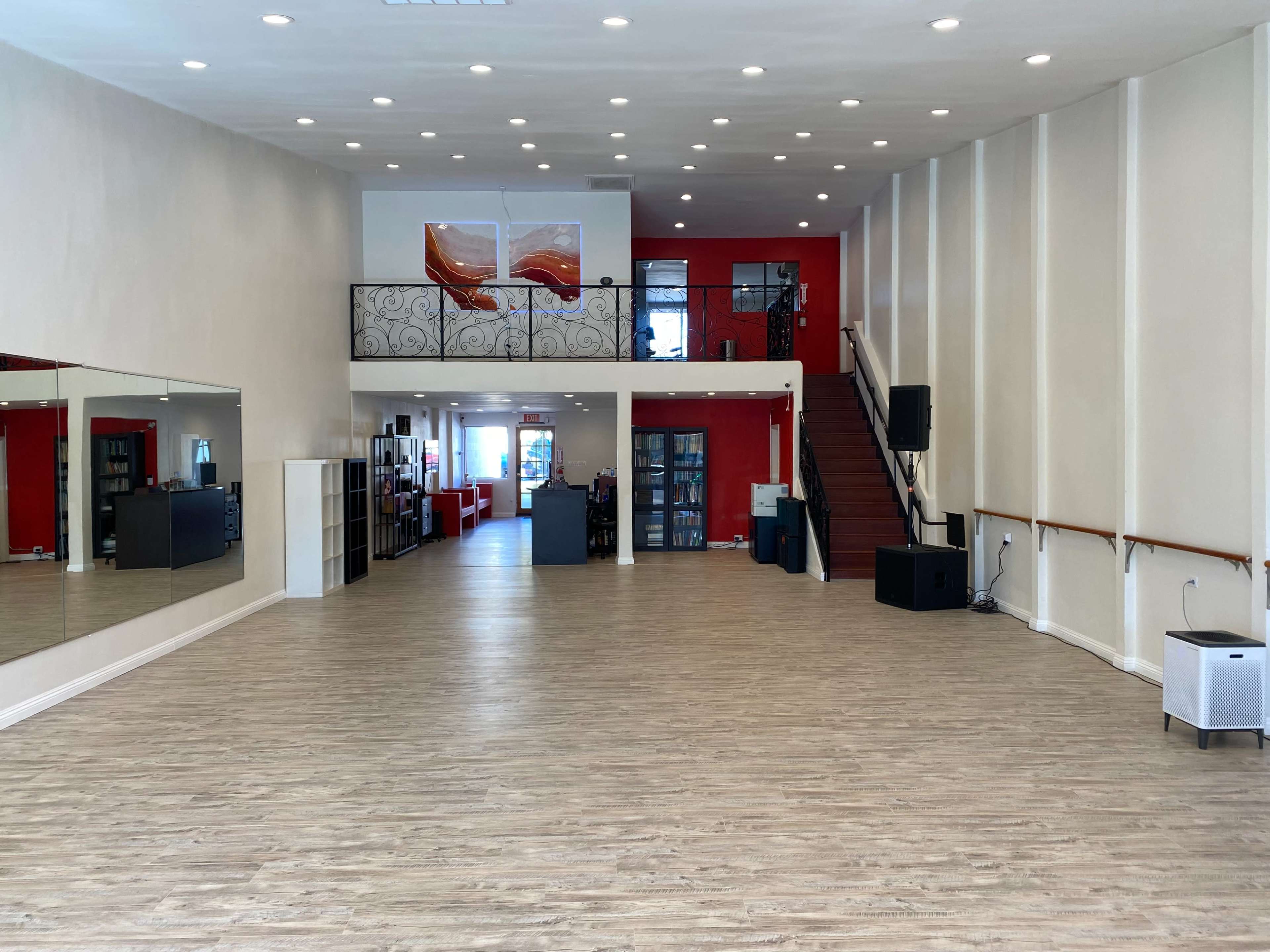 A spacious, well-lit interior of a studio with wooden flooring, white walls, and red accents, featuring a staircase and large mirrors on one side.