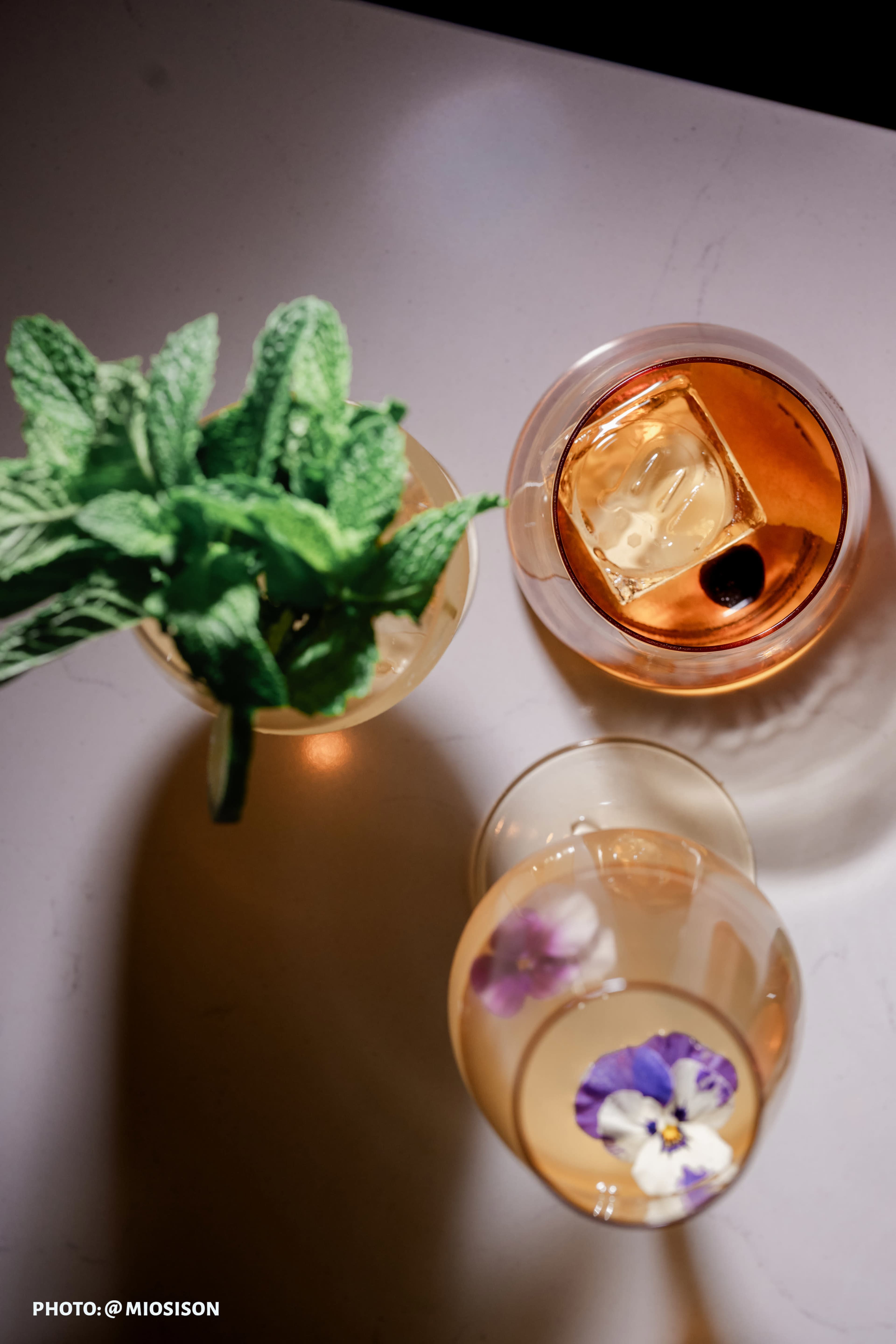 The image shows three glasses of drinks, one with a large ice cube and garnished with mint leaves, and the other two featuring flowers floating on the surface.