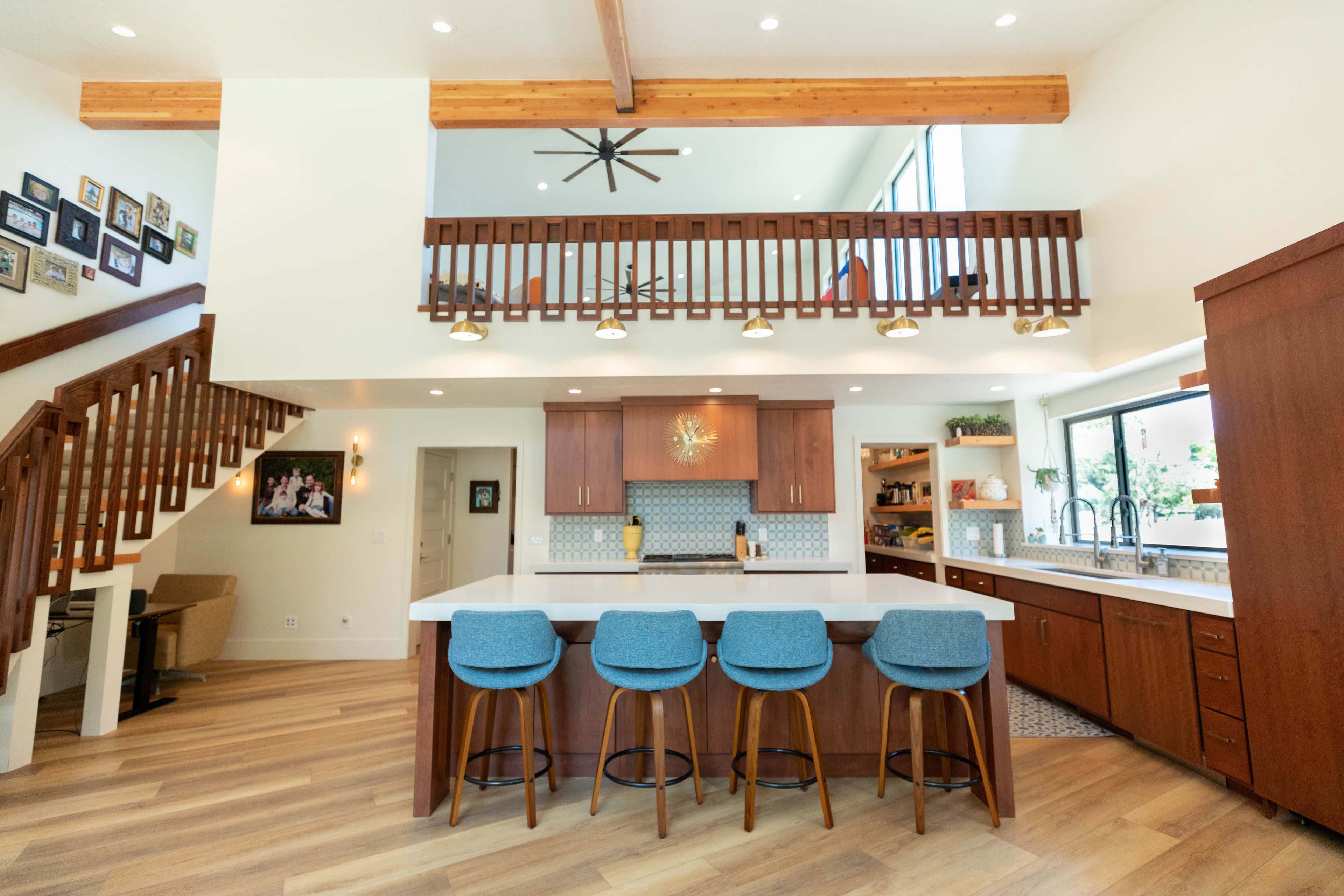 A modern kitchen with a central island and four blue bar stools, complemented by wooden cabinetry and an open design that leads to a balcony above.