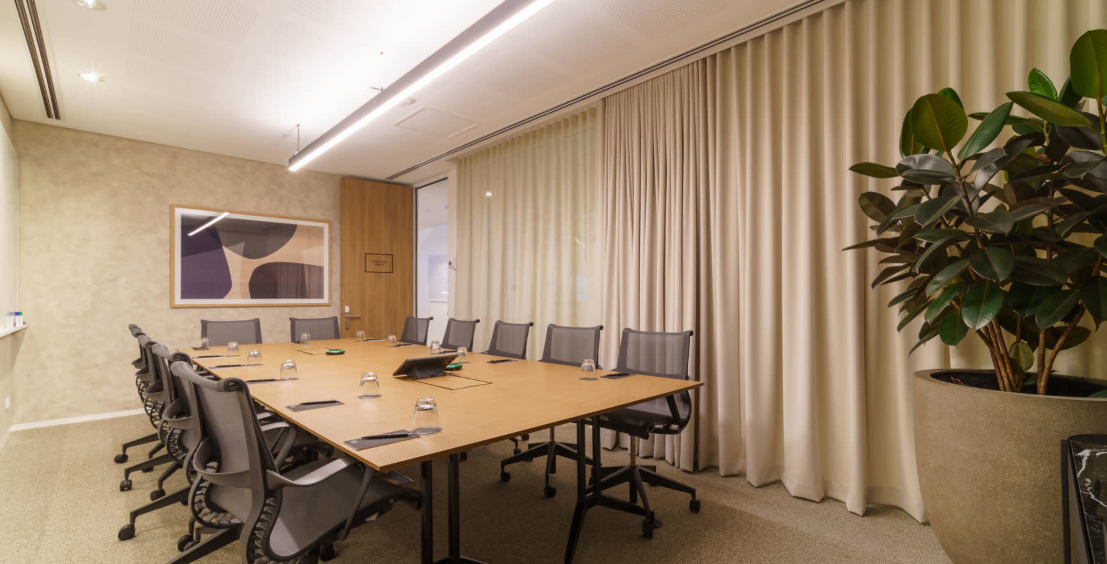 A conference room features a long table surrounded by chairs, with large windows covered by curtains and a plant in the corner.