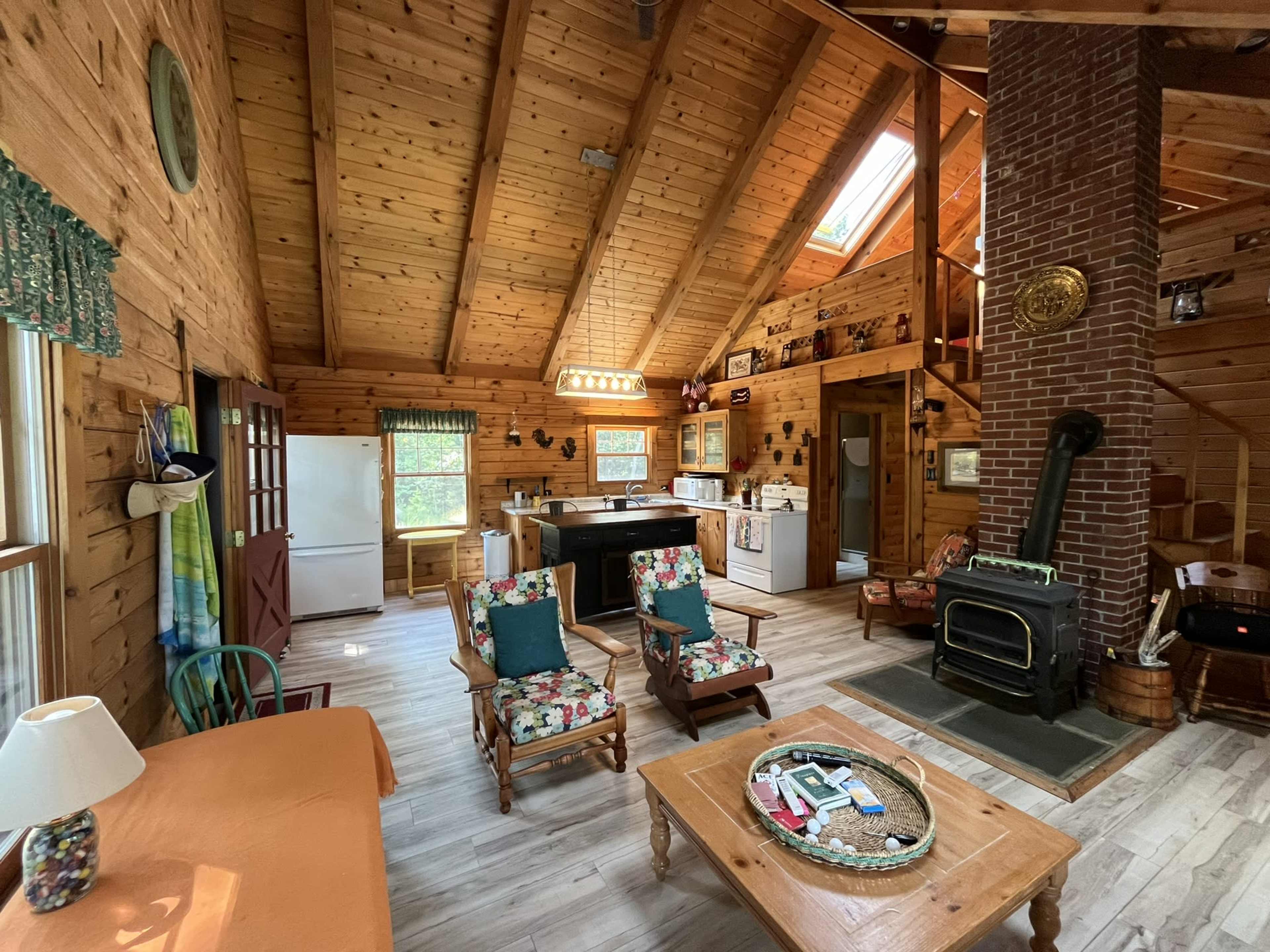 A rustic wooden cabin interior with a kitchen, living area, and a wood stove, featuring furniture arranged around a coffee table.