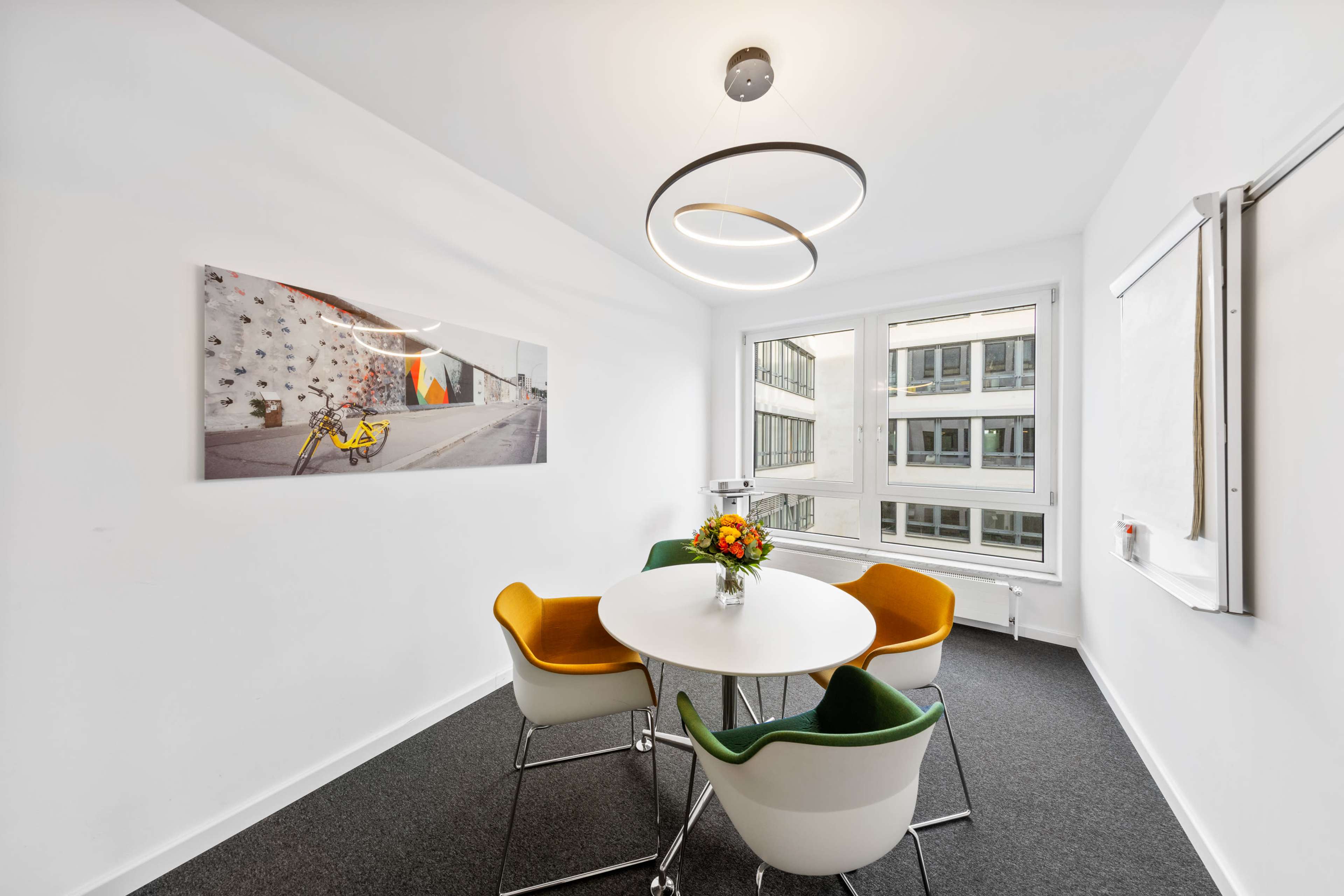 The image shows a bright meeting room with a round table, four colorful chairs, a flower arrangement in the center, and a wall-mounted artwork.