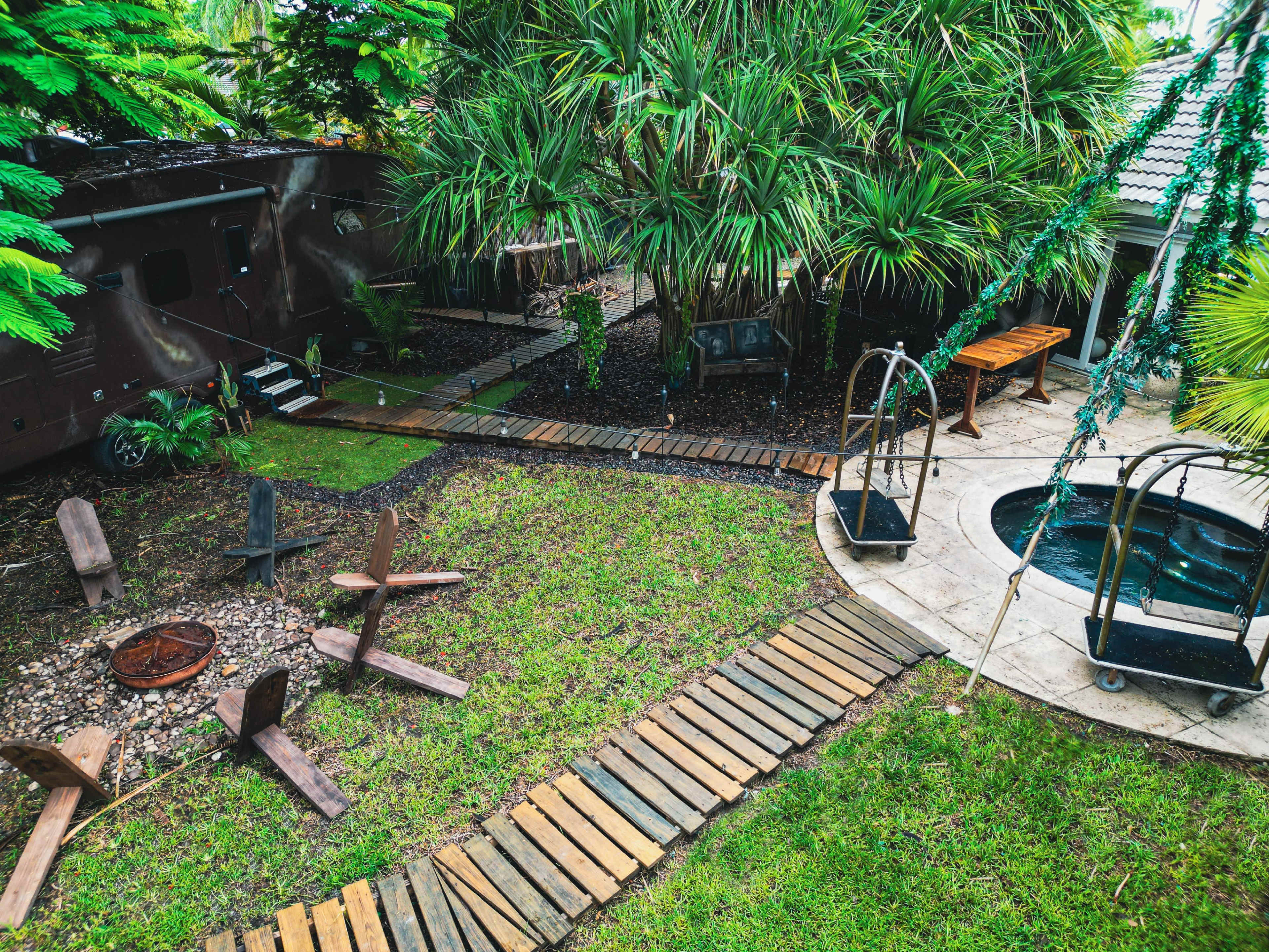 The image shows a landscaped backyard featuring a circular fire pit, wooden pathways, a small pool, and several tropical plants.