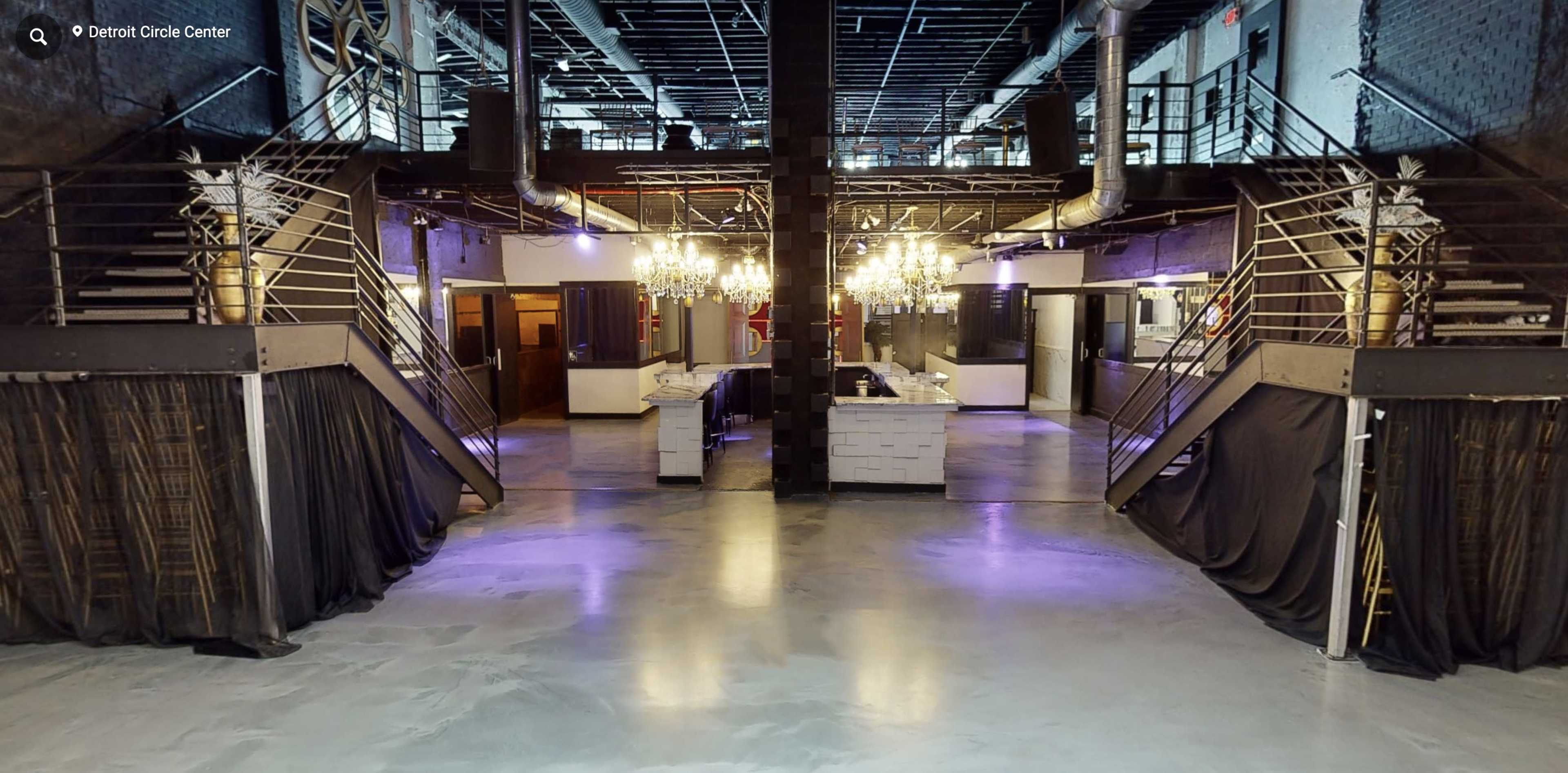 A spacious, modern event venue featuring two staircases leading to an upper level, with a bar area and decorative chandeliers in the center.